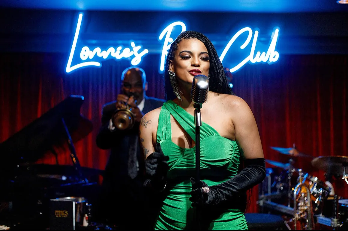 A woman in a green dress singing into a microphone on stage with a jazz band, neon sign background, and a trumpet player.