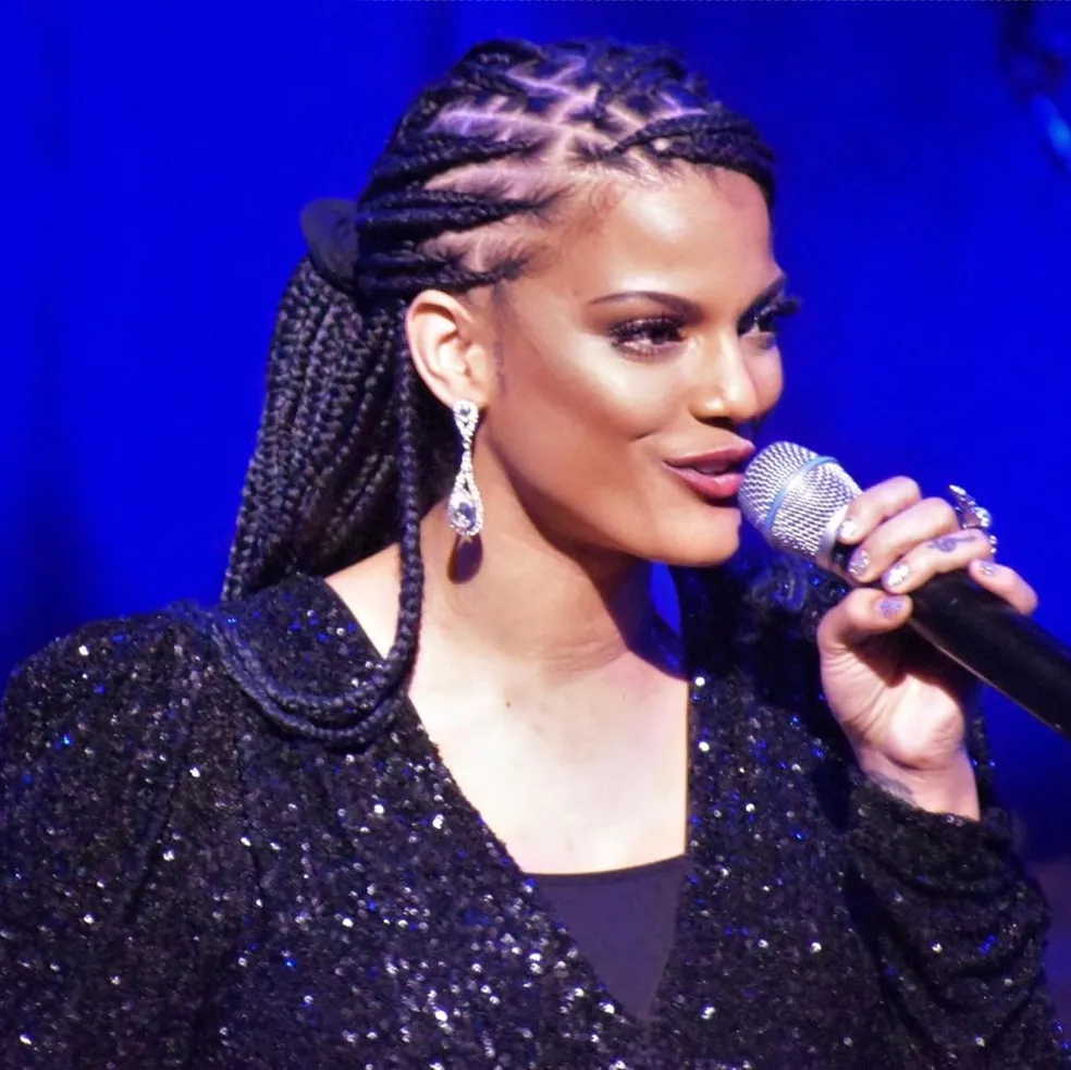 Woman with braided hair and earrings speaking into a microphone, wearing a sparkly black outfit, against a blue background.