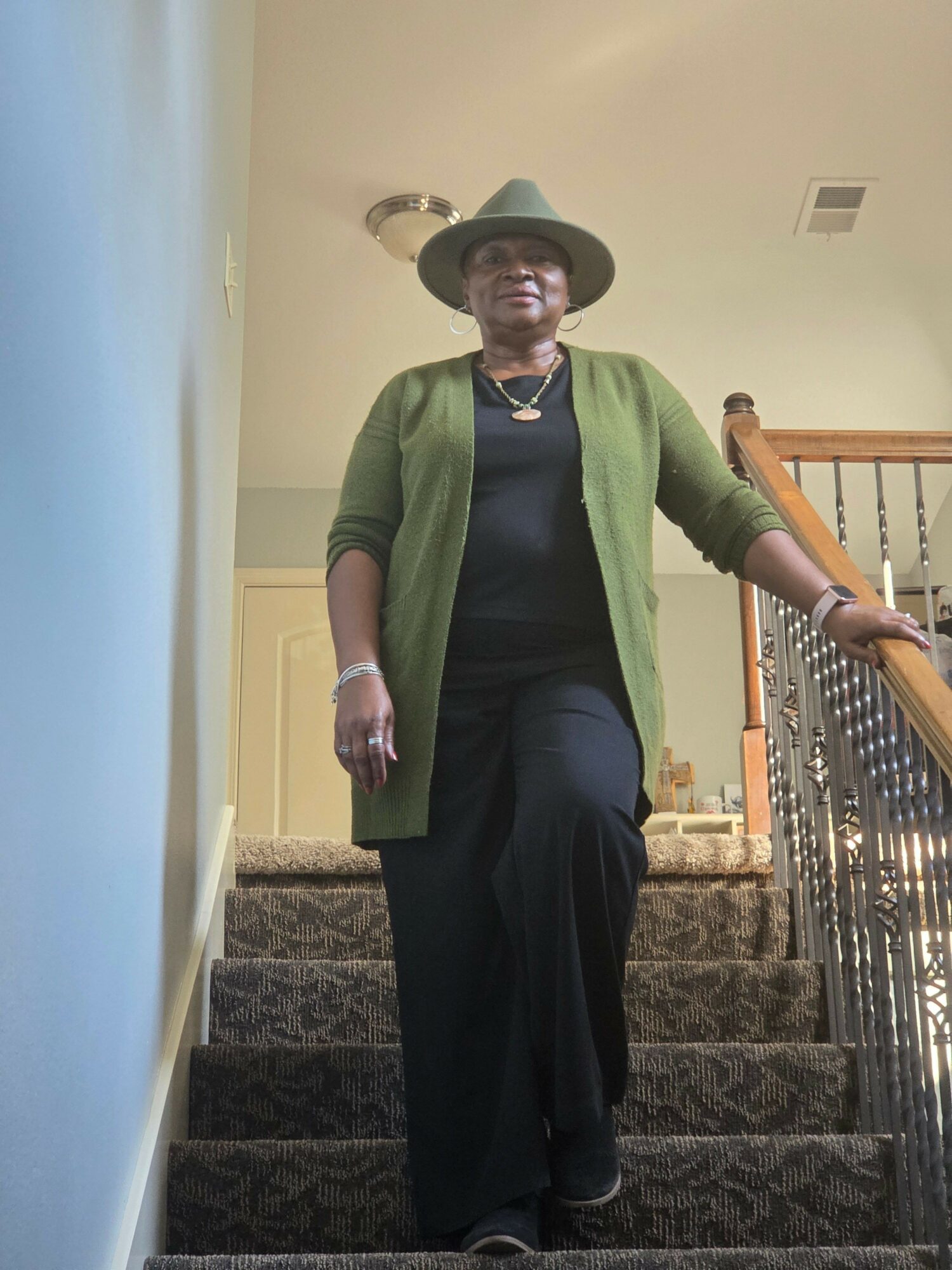Person standing on stairs, wearing a hat, green cardigan, black shirt, and black pants, holding a cane in one hand.