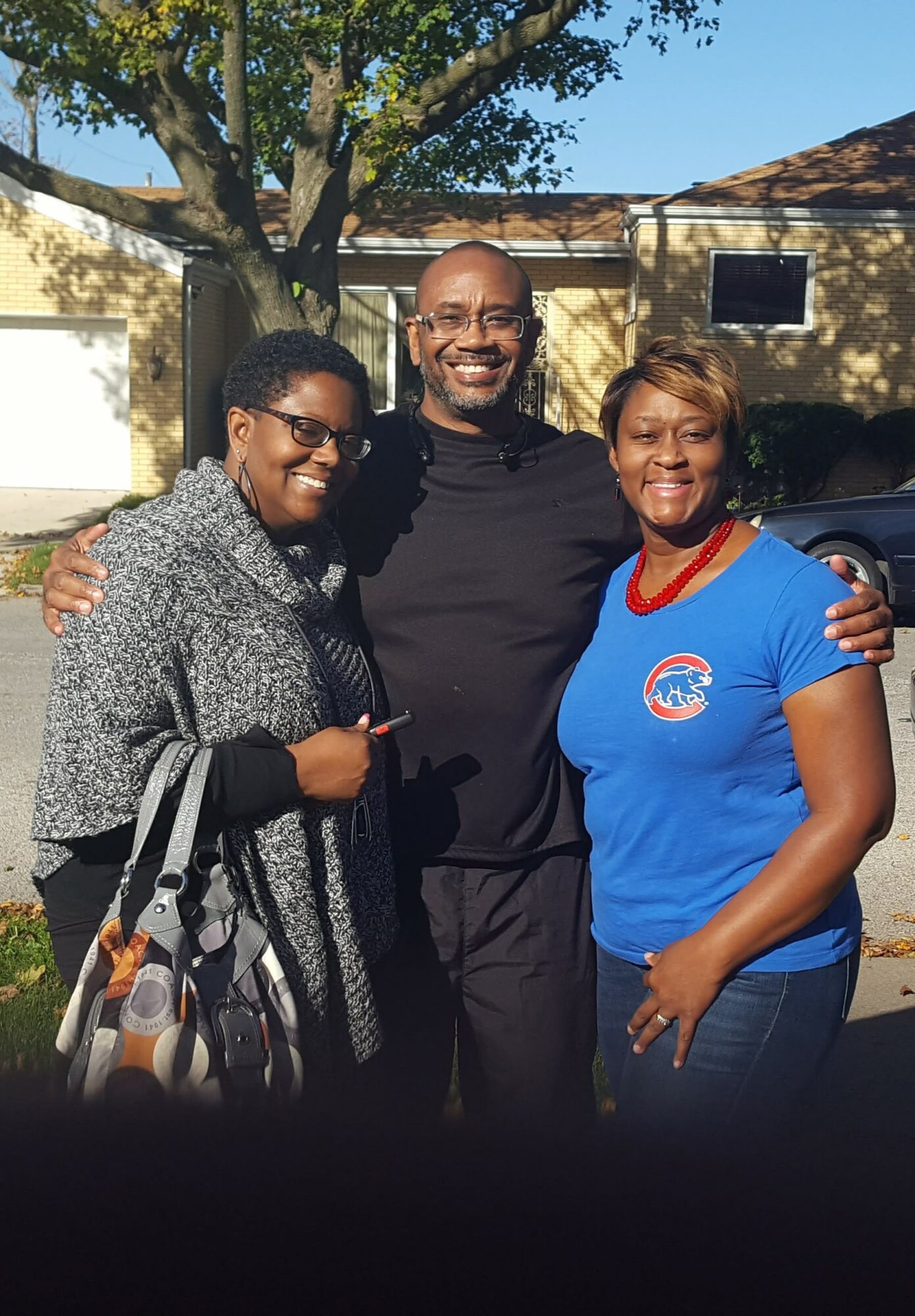 Three people smiling outdoors, standing close together with arms around each other, in front of a tree and building.