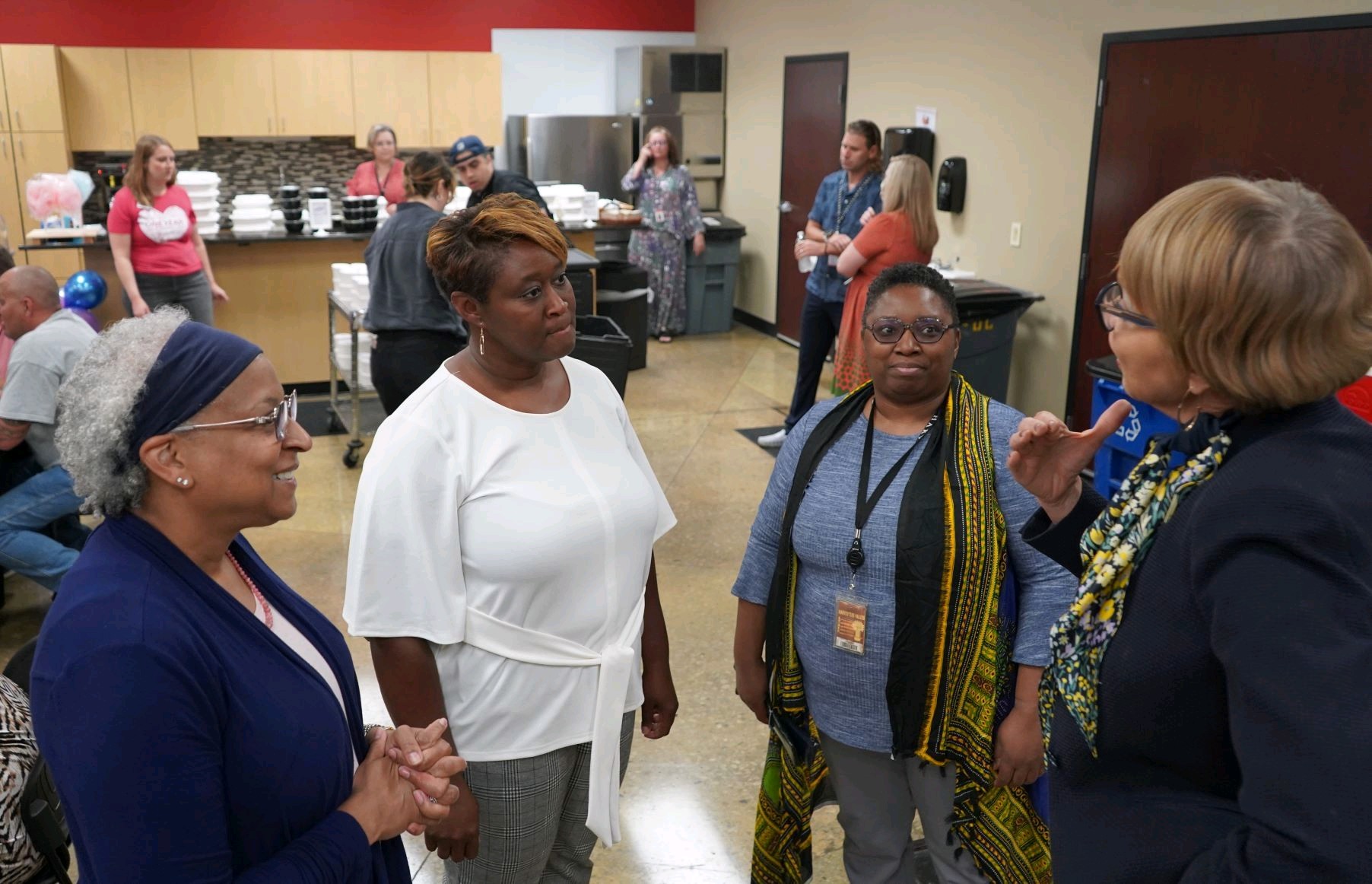 Four women engaged in conversation in a room with people in the background, some near a table with dishes.