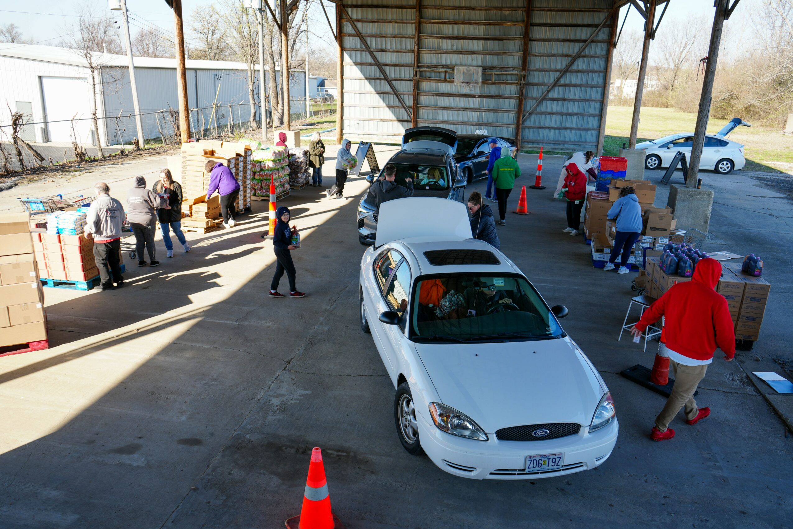 People gather around cars at an outdoor drive-thru or pickup area under a large open-sided shelter, with some people in line.