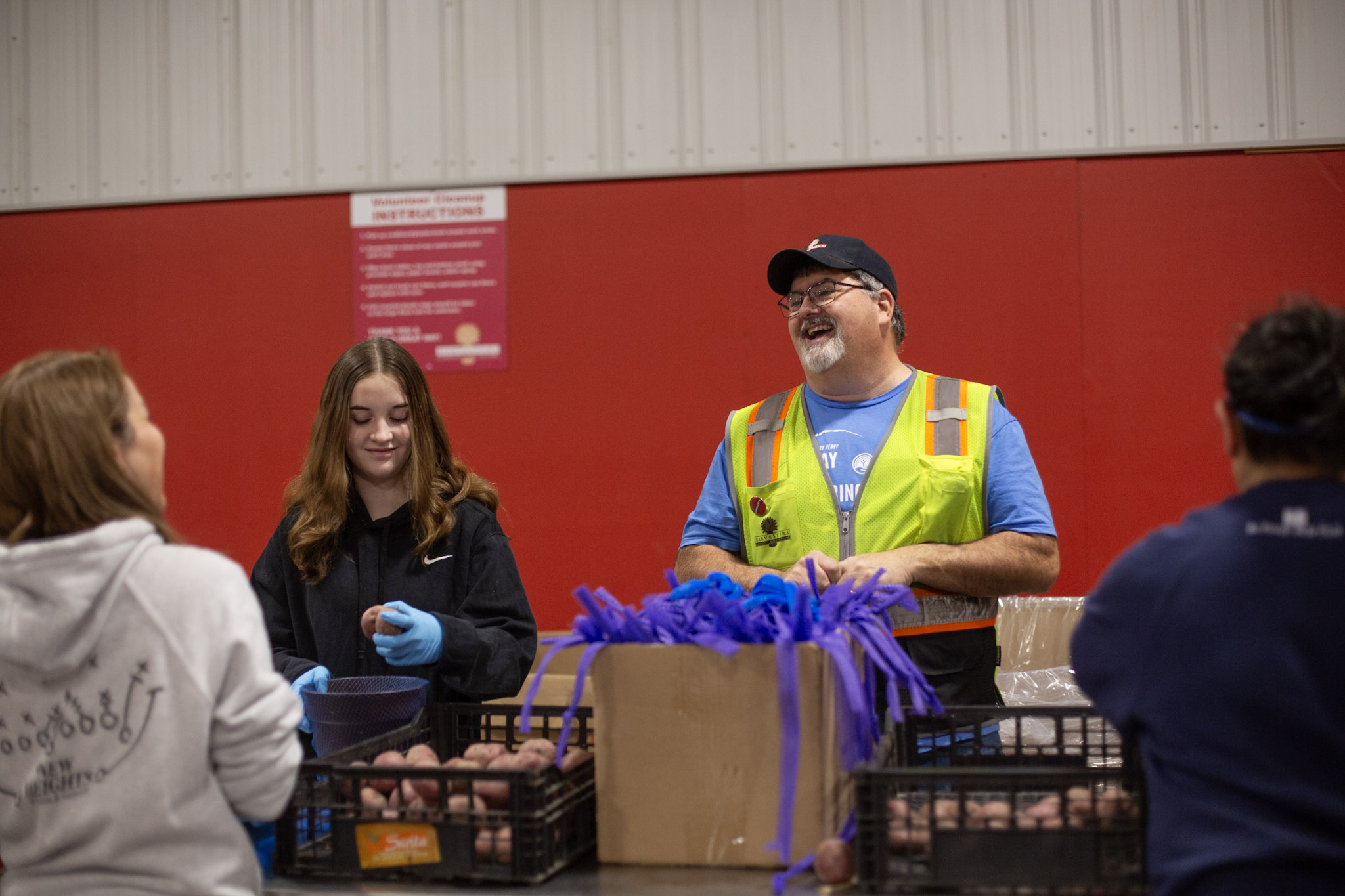 Group of people talking indoors, man in safety vest smiling, woman with gloves, red wall background.