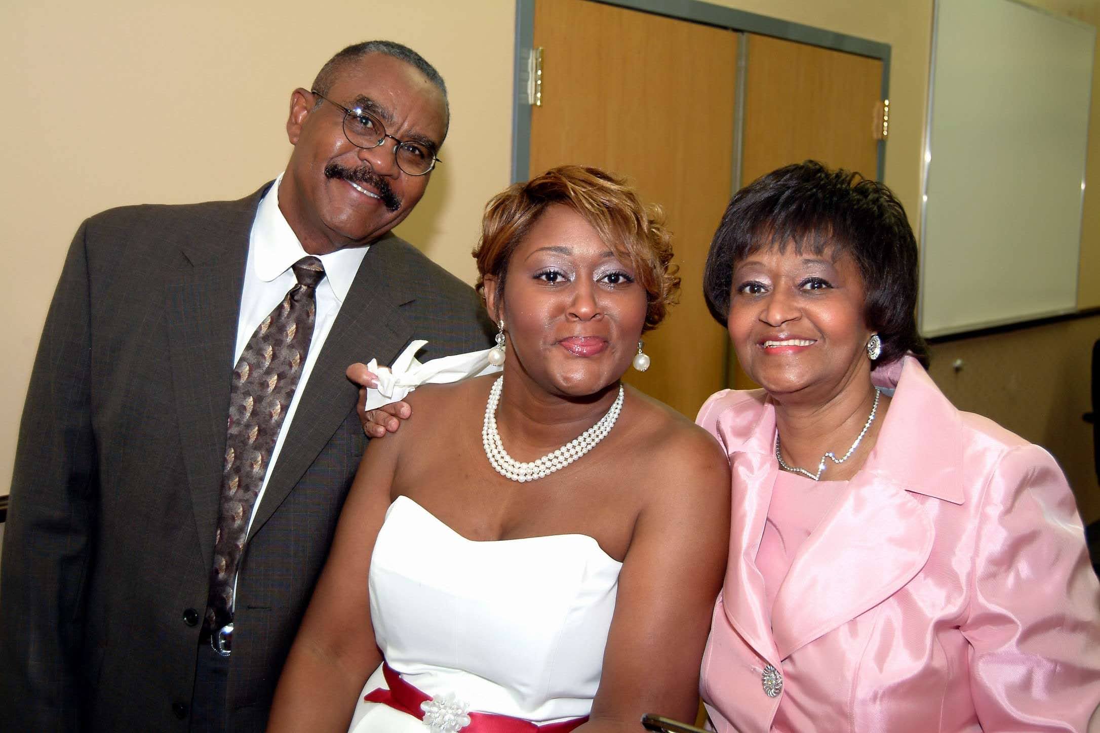 Three people smiling, two women and one man, standing indoors in front of a door and wall, dressed formally.