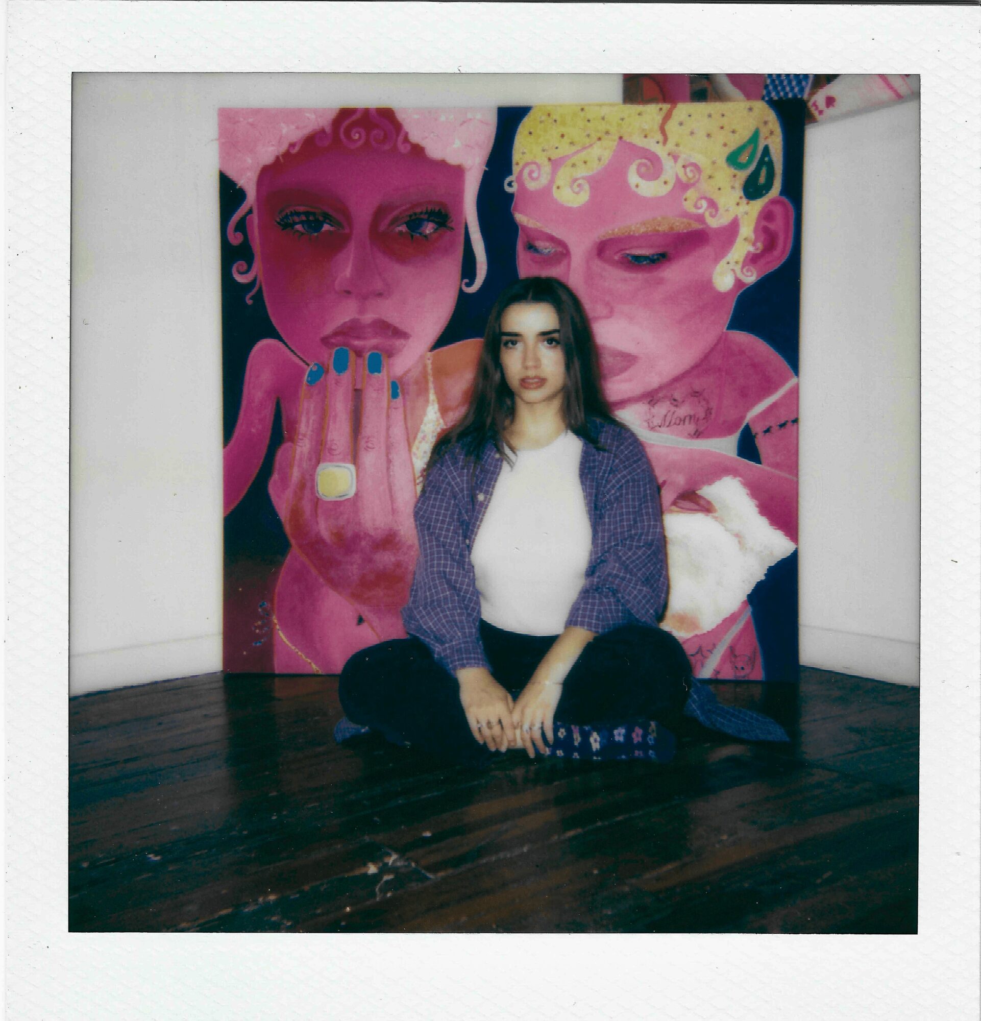 Person sitting on floor in front of colorful mural with two large faces, one pink and one yellow, and abstract designs.