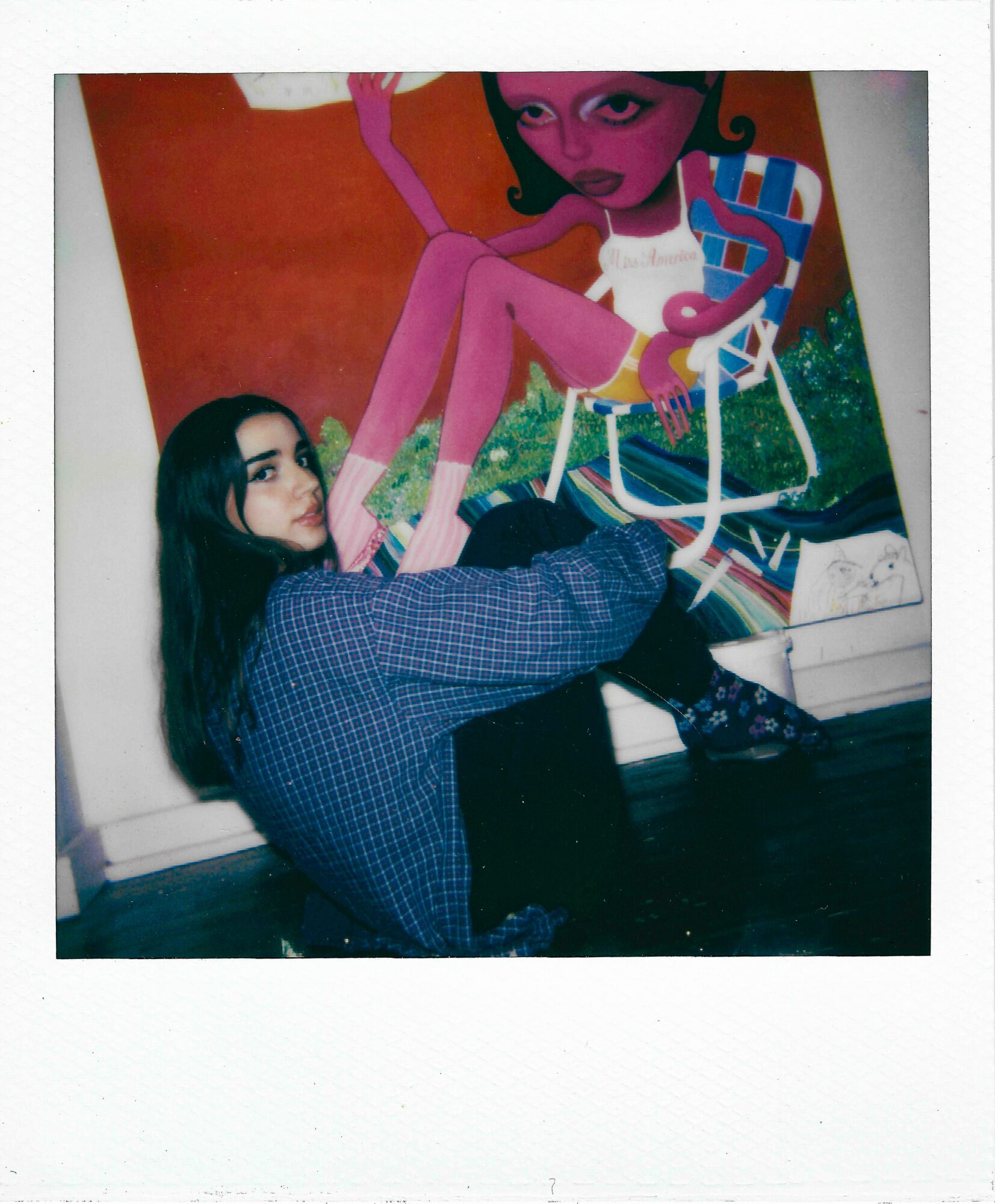 Person with long dark hair sitting on the floor, holding a paintbrush, in front of a colorful mural of a woman with pink skin and green hair.