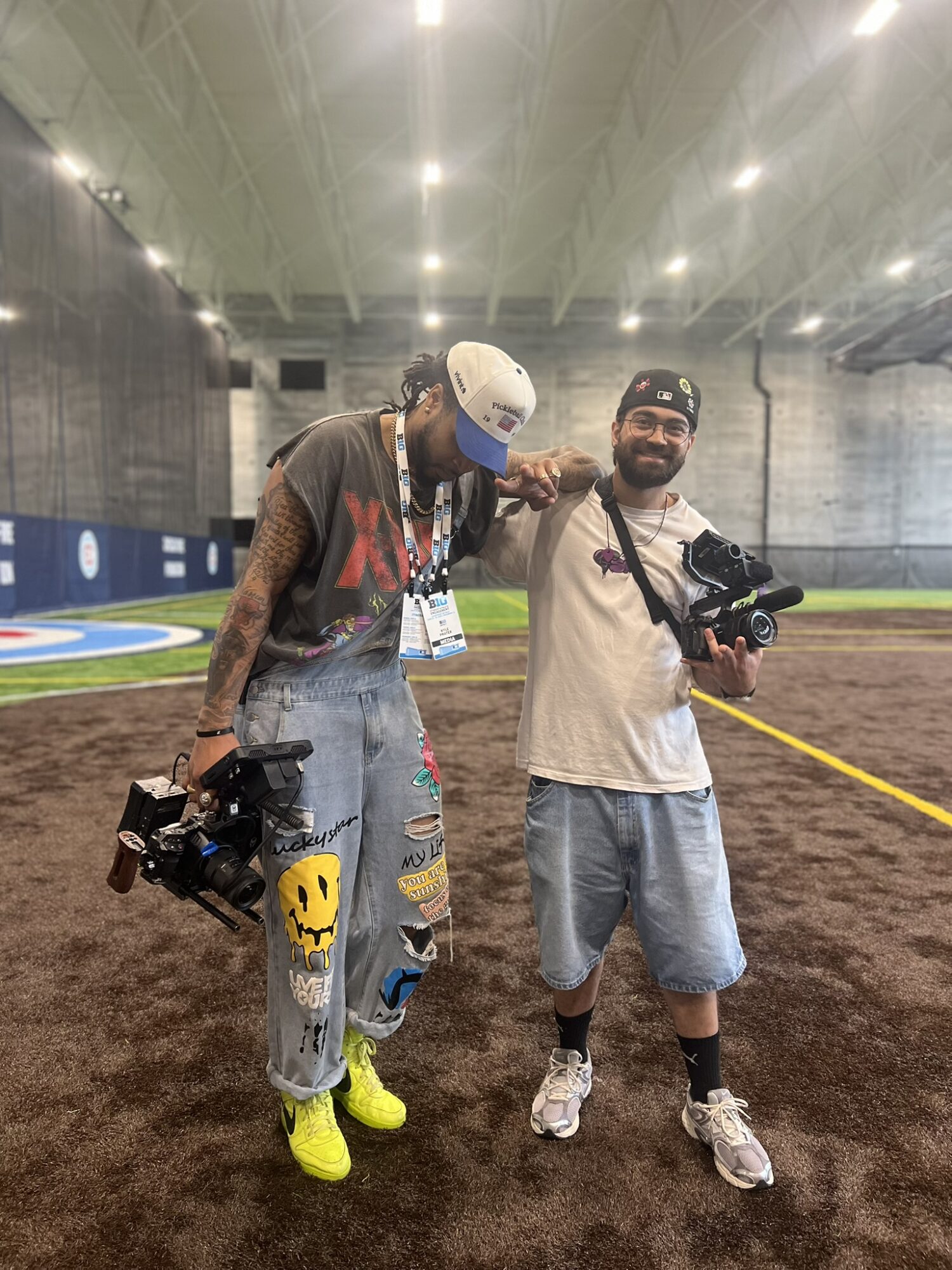 Two men standing inside an indoor sports facility, one holding a camera and the other with a camera on his shoulder, smiling.