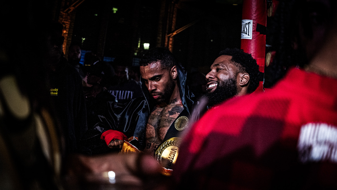 Two men with muscular builds and tattoos, one smiling, in a dark environment with red and black clothing, possibly a boxing event.