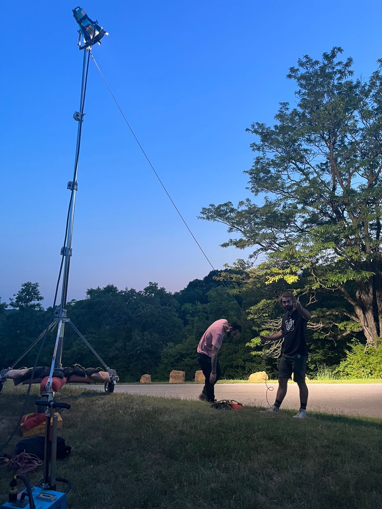 Two people standing outdoors near a tree, with a tall lighting or camera tower on the left, under a clear blue sky.