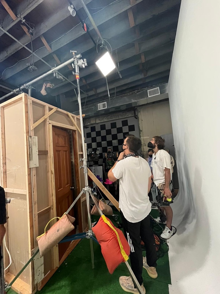 Two people in a studio or set, one with a camera, standing near a wooden door frame and a white wall.