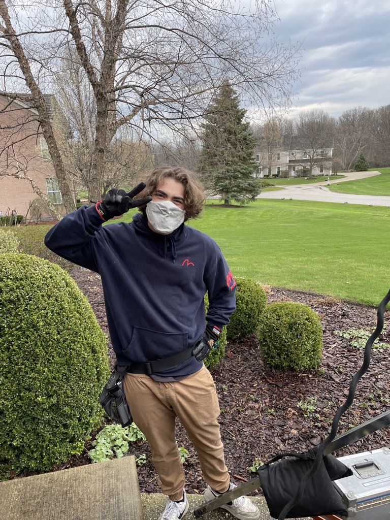 Person wearing a face mask, glasses, navy jacket, and tan pants, standing outdoors near bushes and trees, making a peace sign.