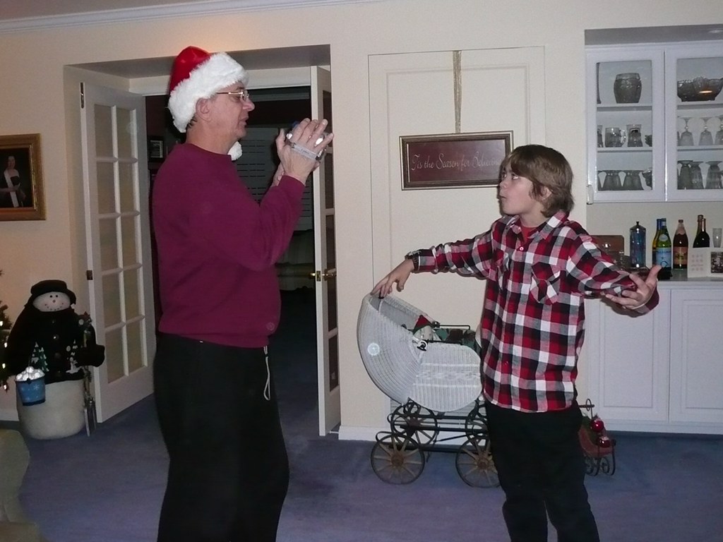 Two people, one wearing a Santa hat, are indoors with a white cabinet and decorations in the background.