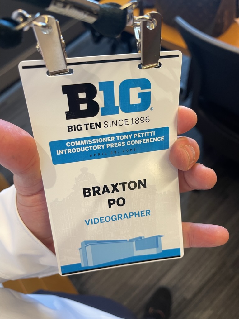 Hand holding conference badge with blue and black text, logo, and a blue graphic at the bottom.