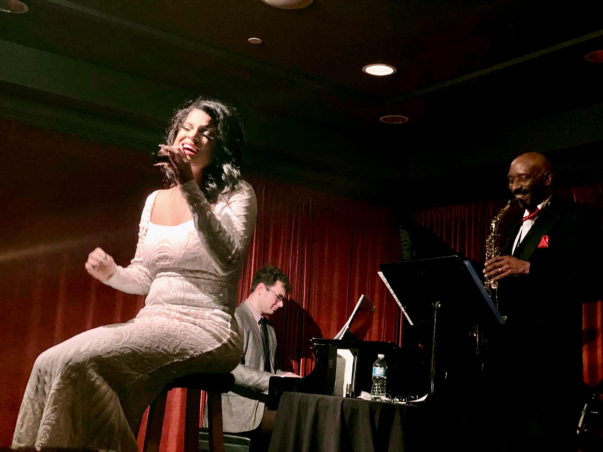 A woman in a white dress sings with a pianist and a saxophonist on stage, red curtains in the background.