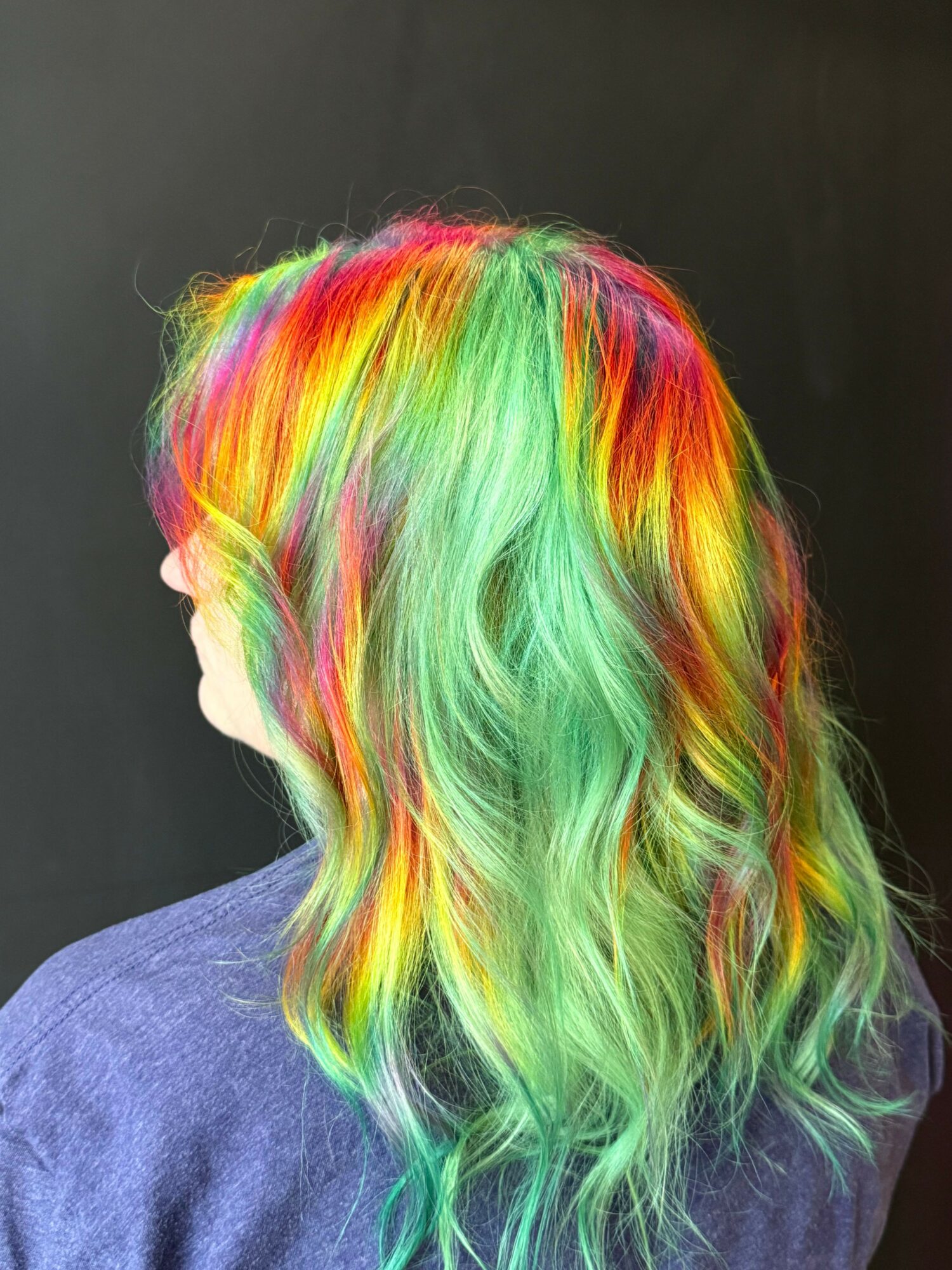 Person with long, wavy hair dyed in rainbow colors, facing downward, wearing a dark top, against a dark background.