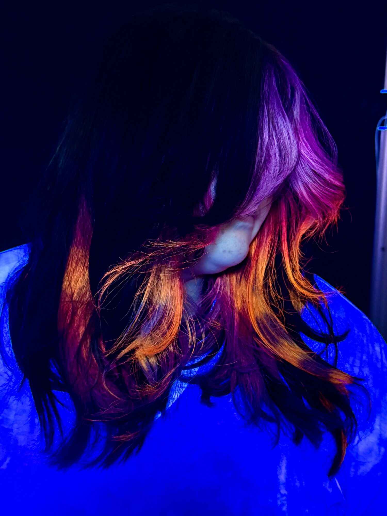 Person with colorful hair looking downward, illuminated by blue and purple lighting.