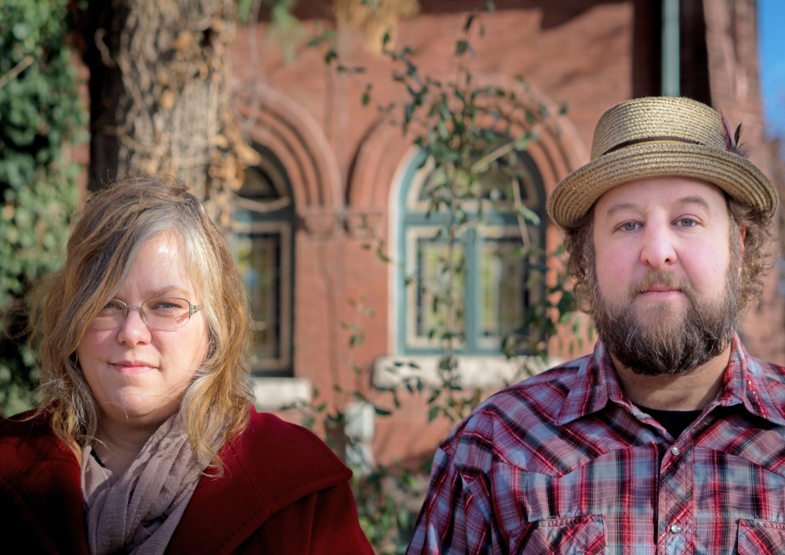 Two people outdoors in front of a brick building with arched windows, one wearing glasses and a red coat, the other with a hat and plaid shirt.
