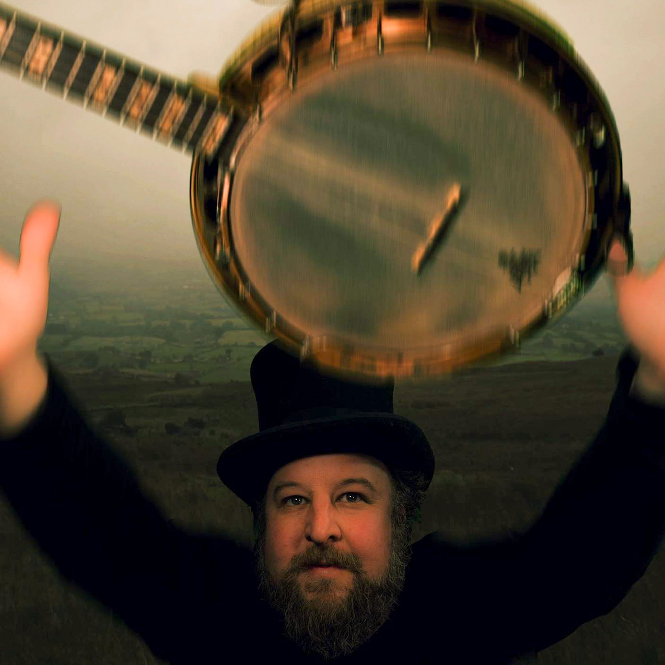 Man with a beard and hat holding a banjo overhead outdoors at sunset.