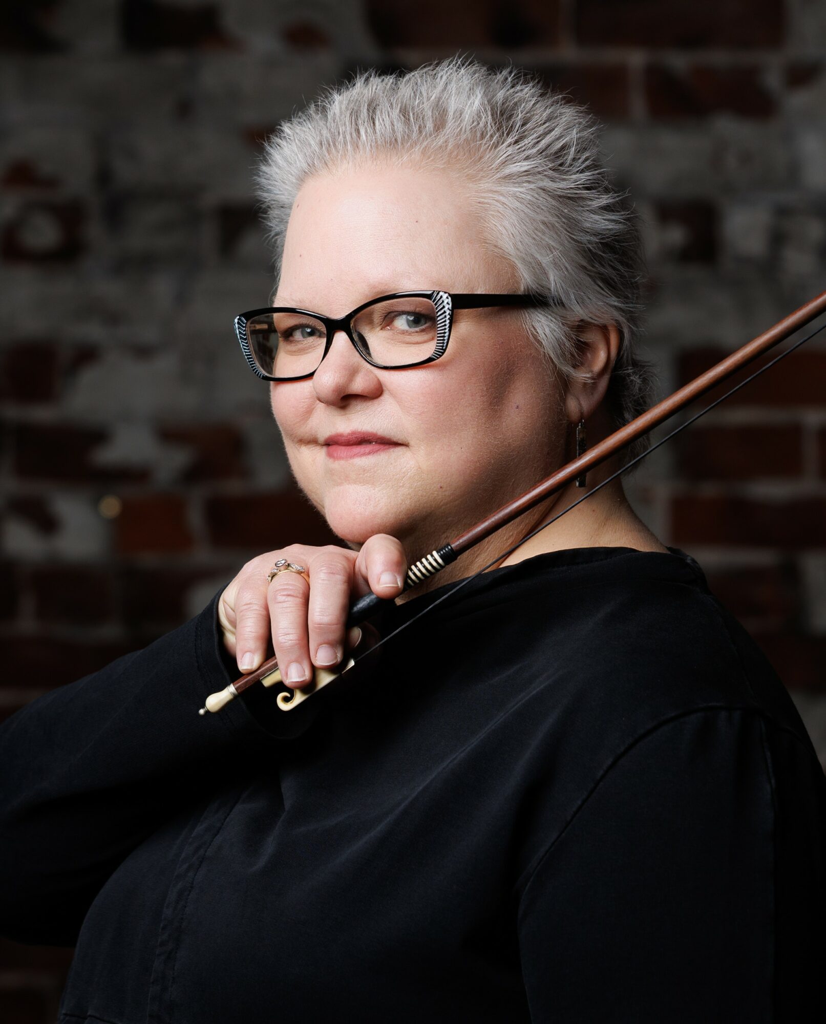 Older woman with gray hair and glasses holding a violin bow, looking at the camera, against a brick wall background.