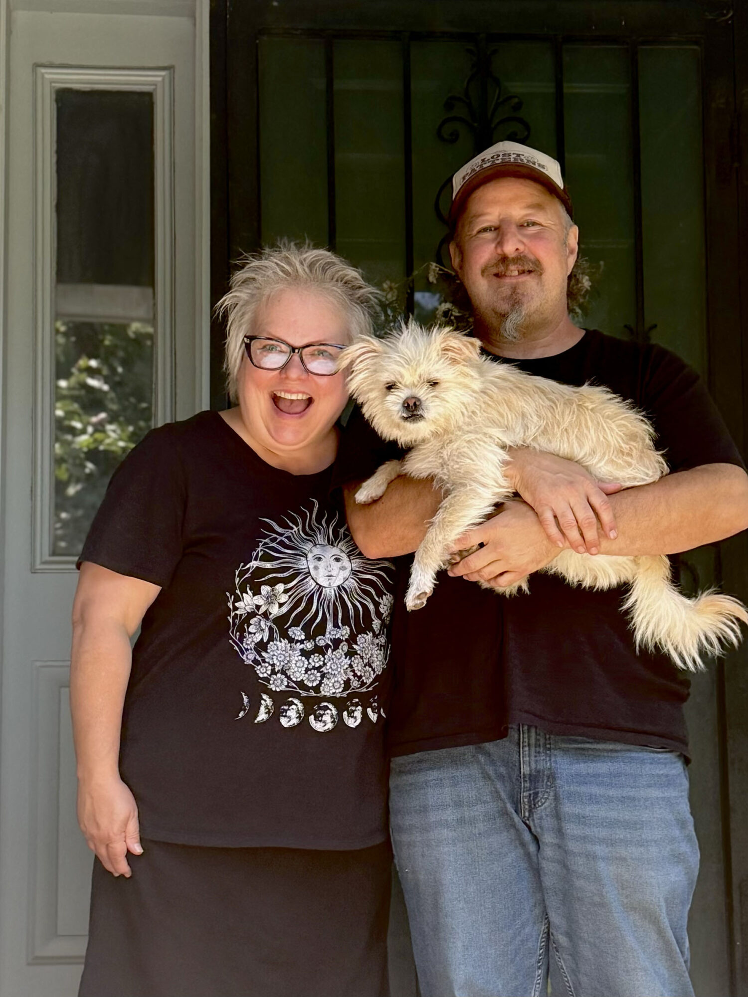Two people smiling, one holding a small dog, standing outside a house with a window and door behind them.