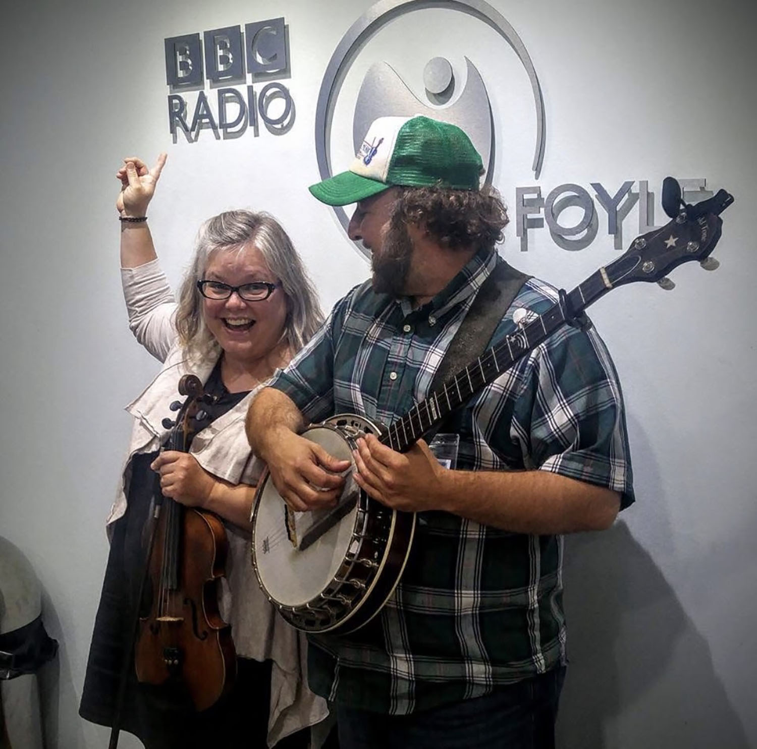 Two people smiling, one holding a violin and the other playing a banjo, in front of a wall with BBC Radio logos.