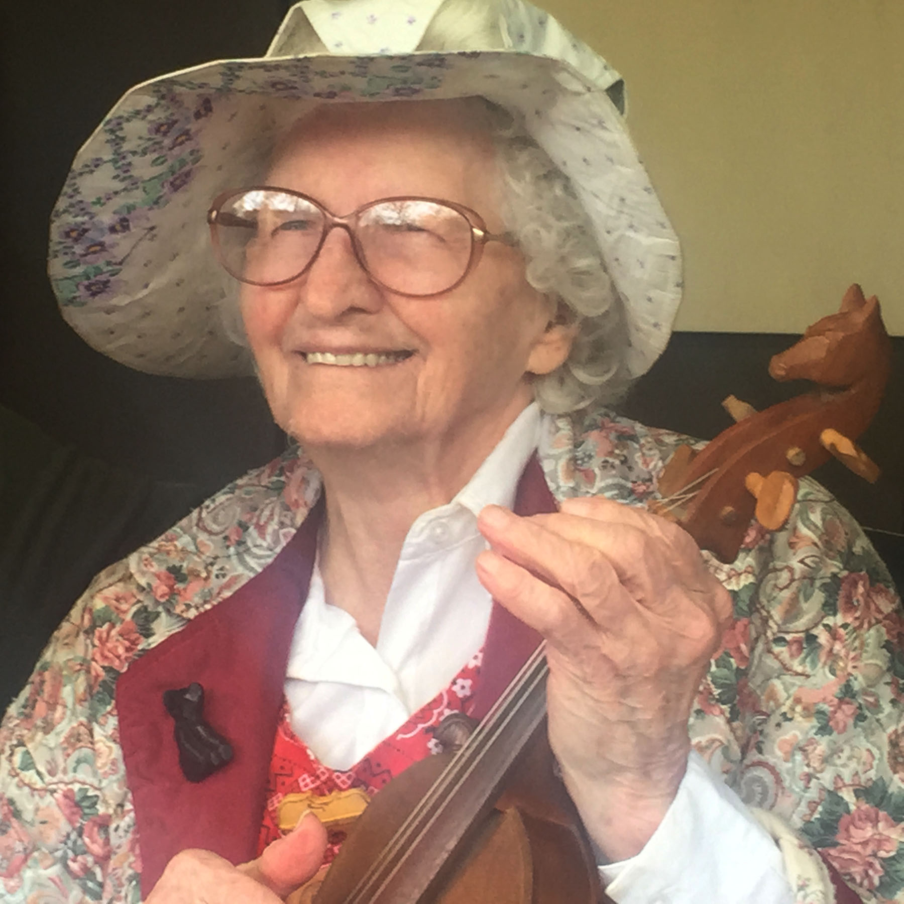 Older woman smiling, wearing glasses, a floral hat, and holding a violin, with a decorative background.