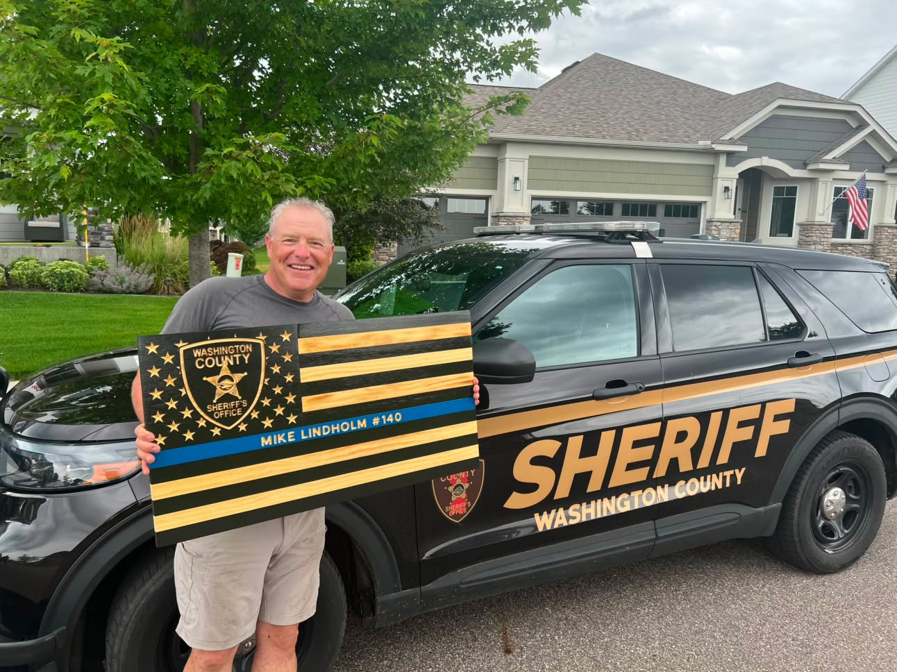 Man smiling holding a sheriff-themed sign in front of a black sheriff vehicle parked on a residential street.