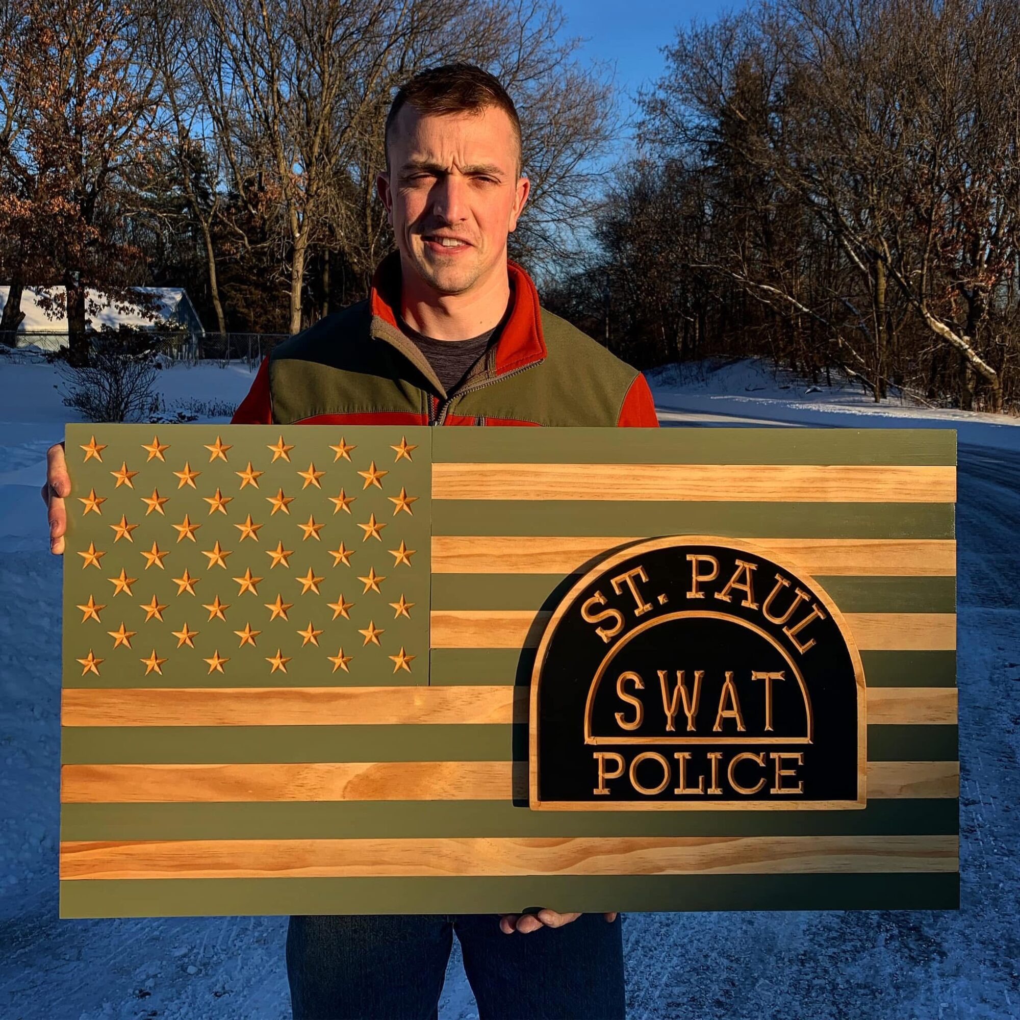 Person holding a wooden American flag with a SWAT police emblem, outdoors with trees and snow, sunny day.