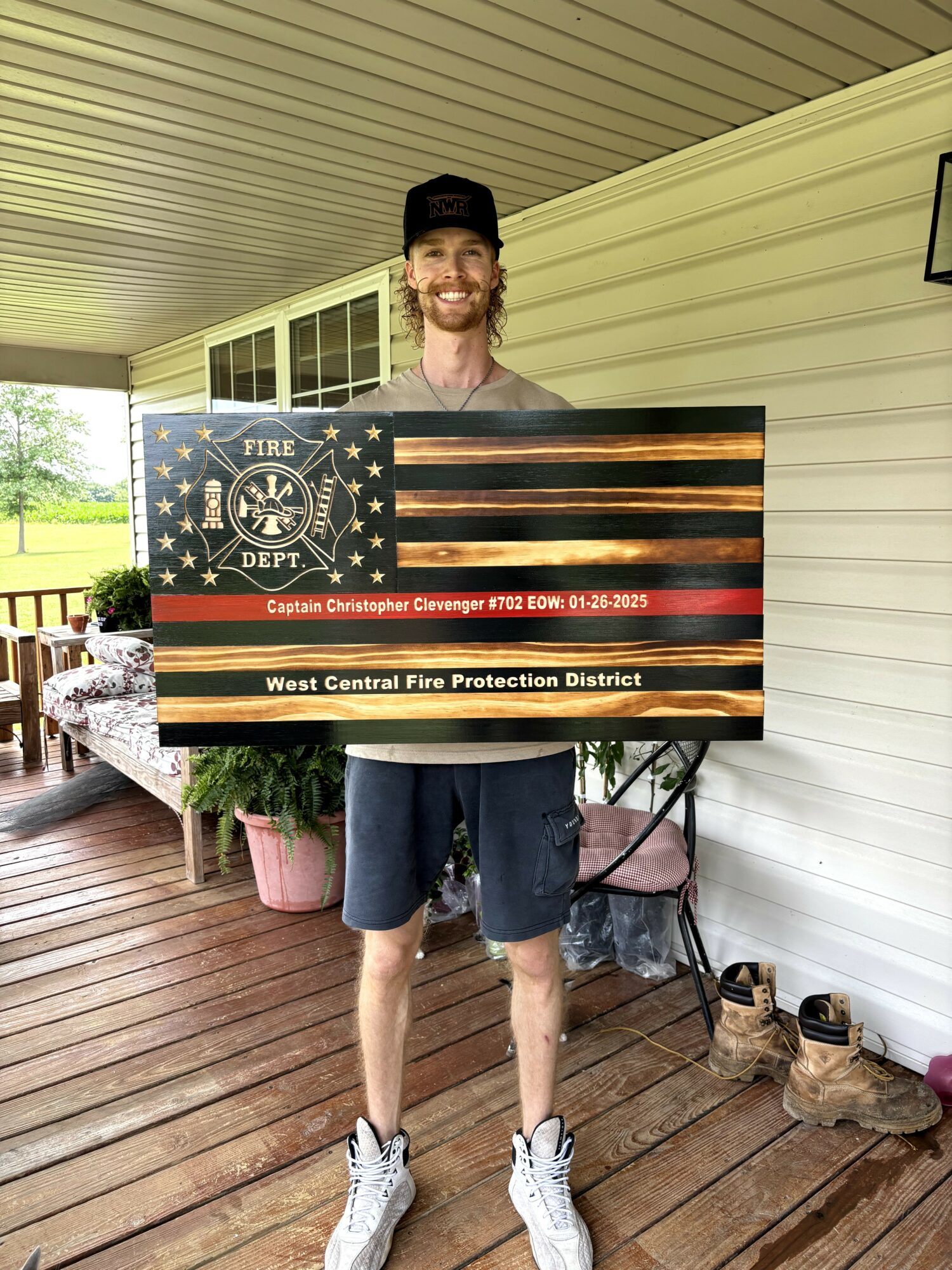 Young man with a beard and cap holding a fire department sign on a porch, smiling, with shoes and plants nearby.