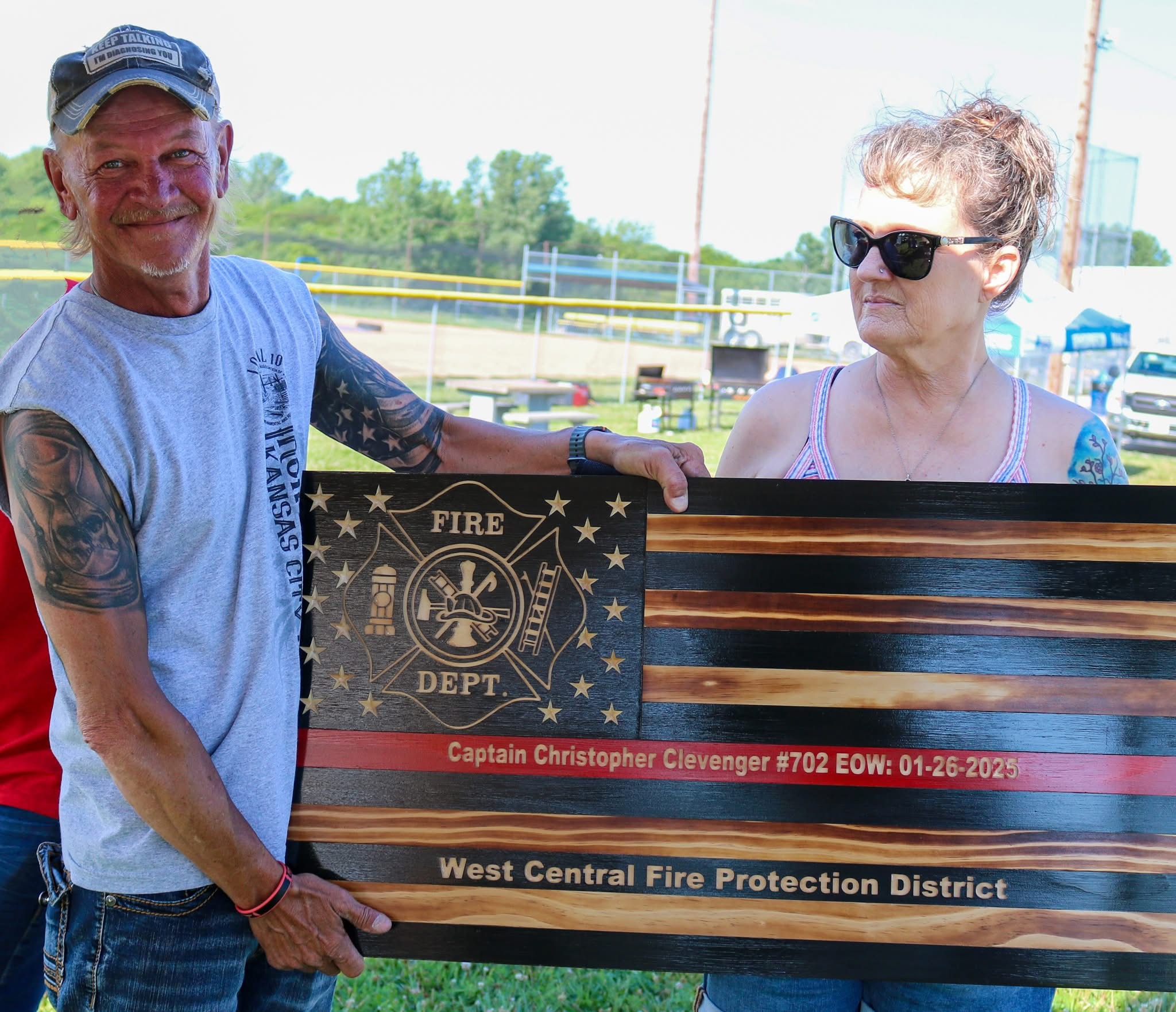 Two people holding a flag-shaped sign outdoors, with a fence and trees in the background.