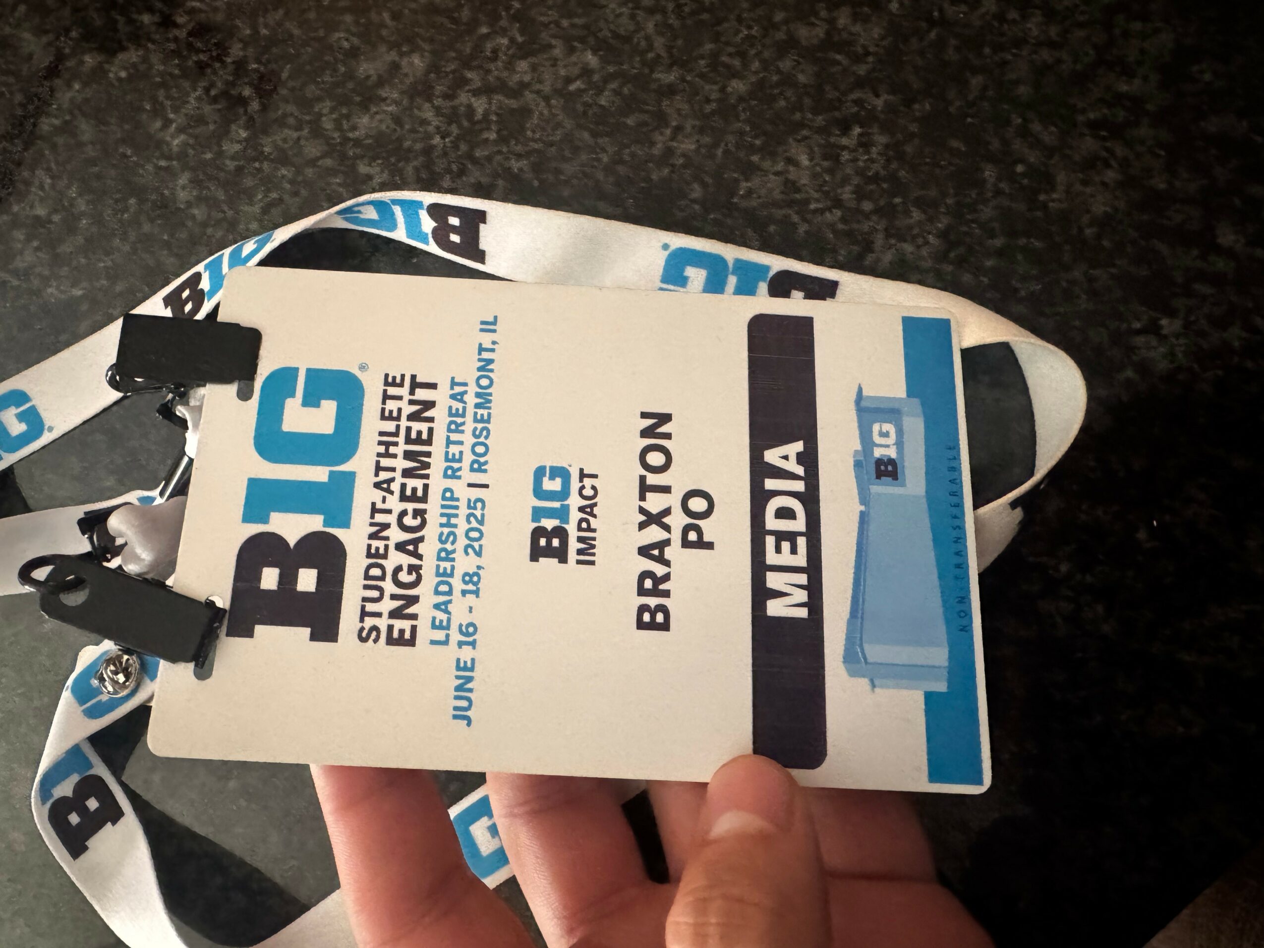 Event badge with blue and white design, held by a person's hand, displaying text about a media event, on a dark surface.