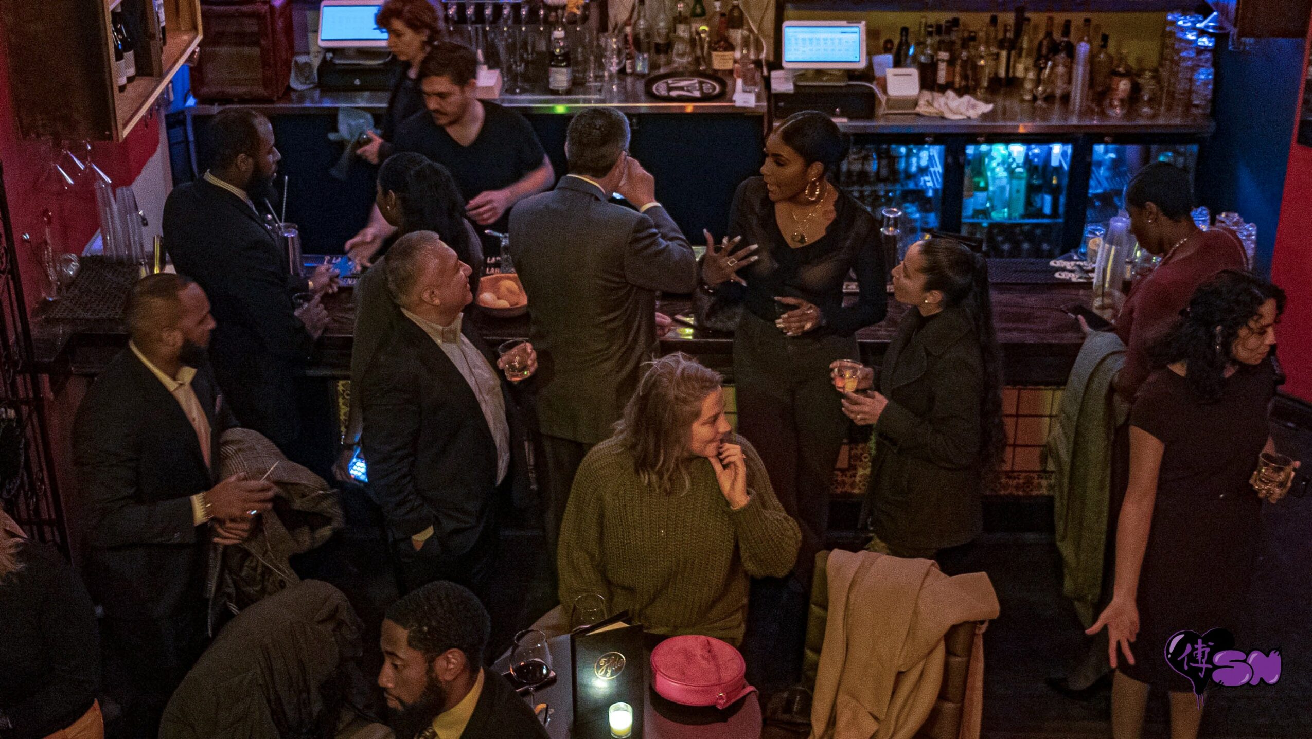 People socializing and drinking at a bar with a bartender in the background, some standing and some seated.