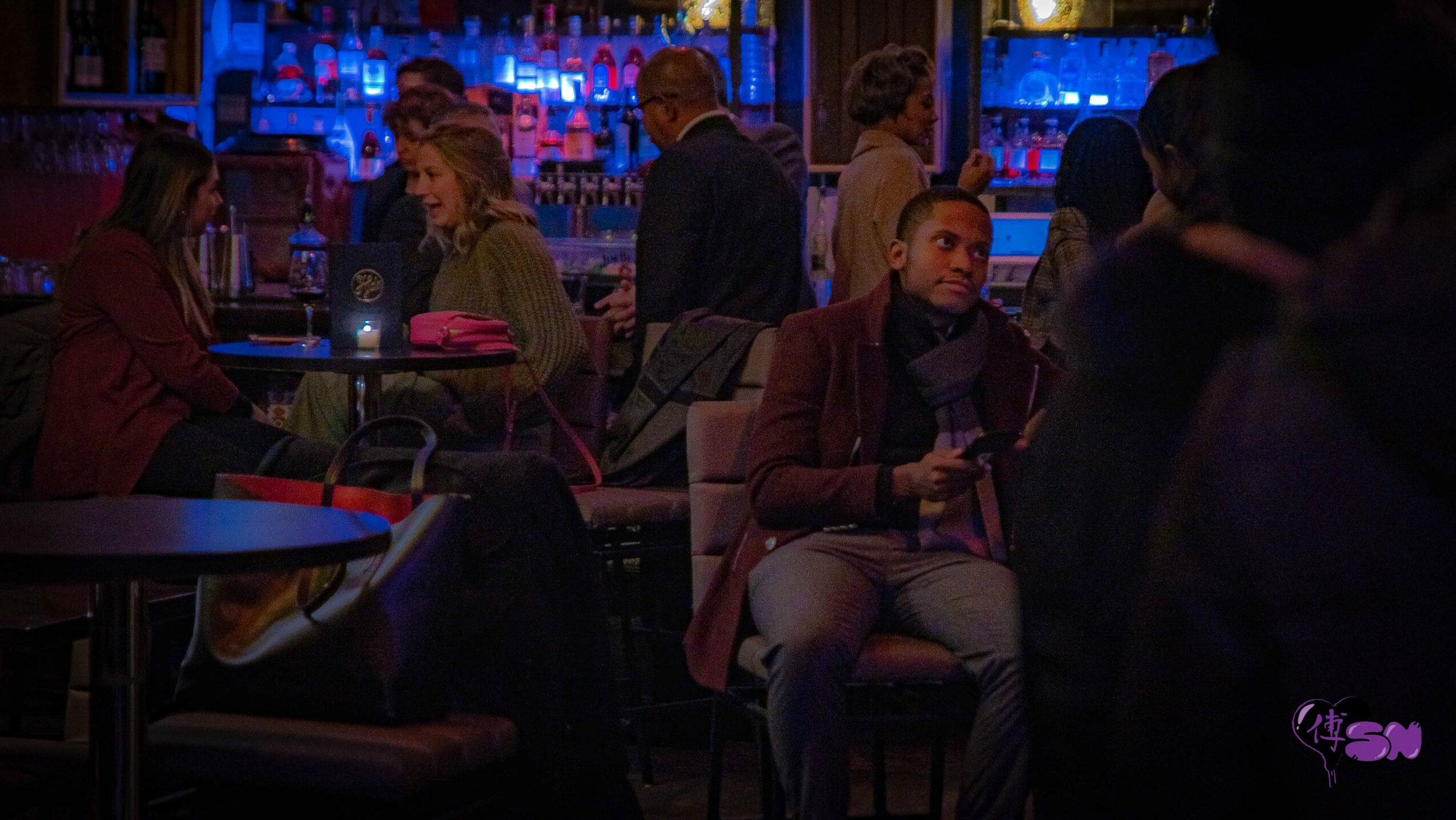 People sitting and socializing in a dimly lit bar with blue and purple lighting, some holding drinks, background shelves with bottles.