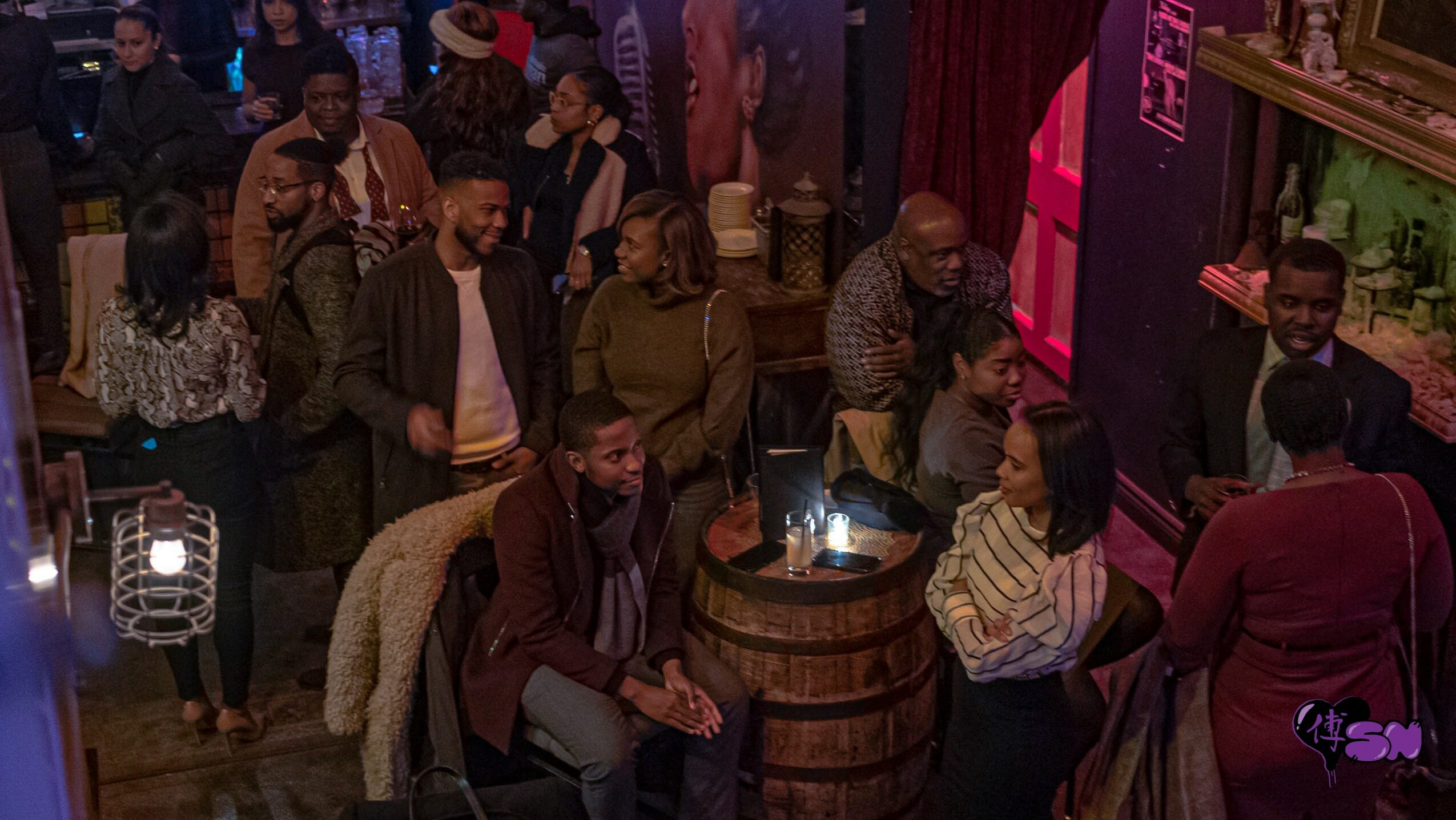 People socializing in a dimly lit bar or club with a wooden barrel table, some standing and some sitting, colorful lighting.