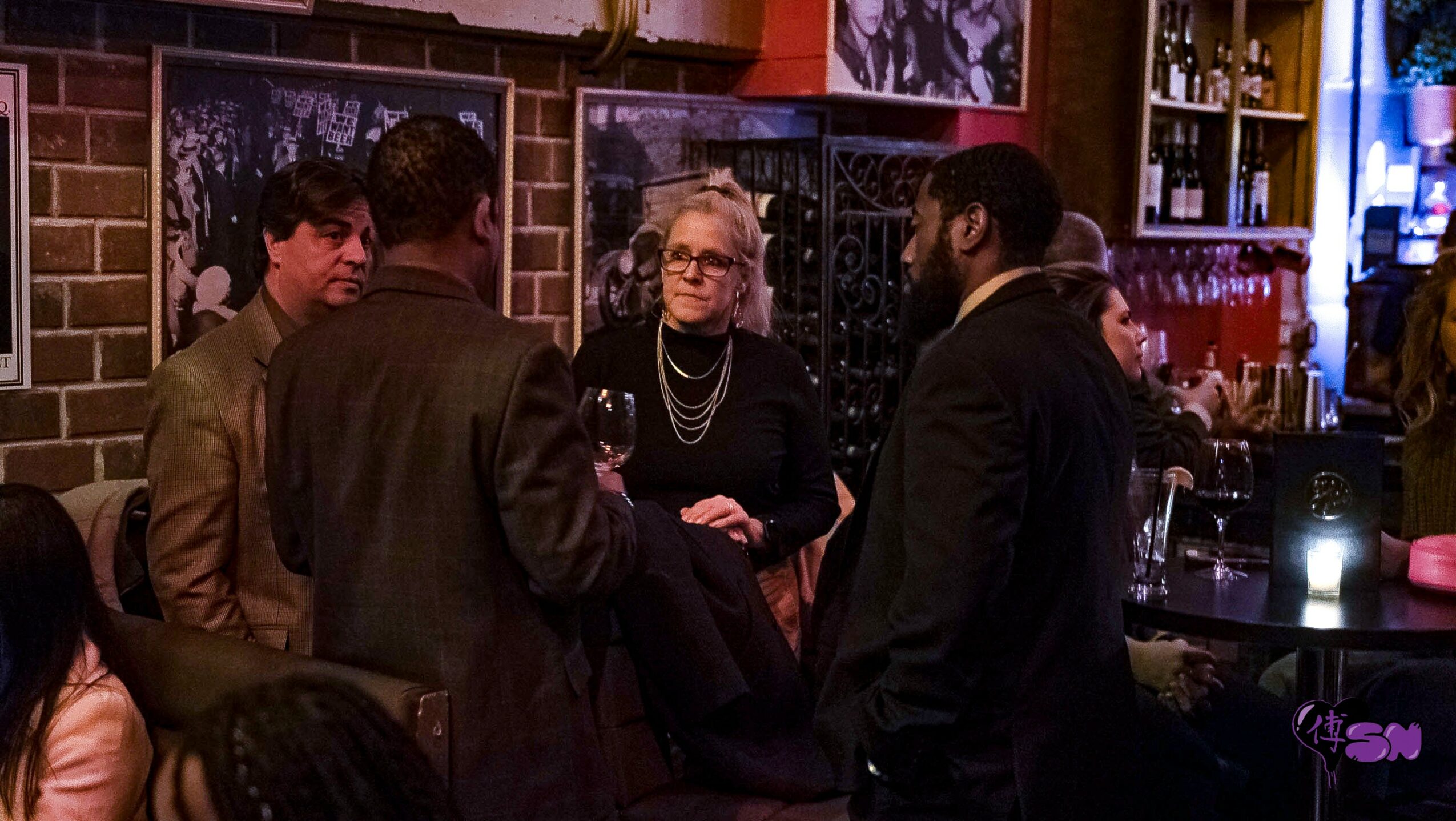 Group of four people talking in a dimly lit bar or restaurant, with brick walls and framed photos in the background.