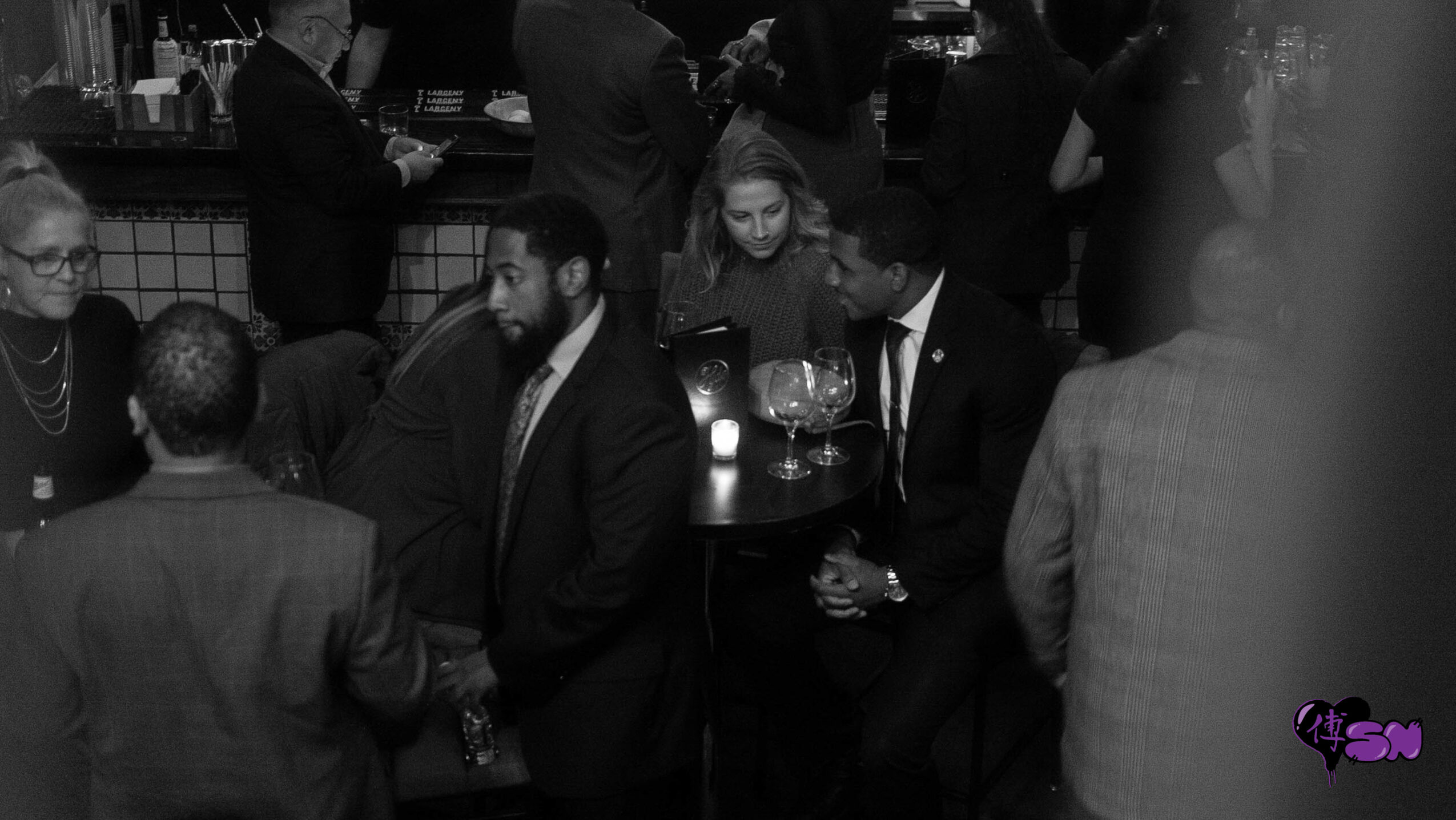 People gathered around a table at a social event, some holding drinks, in a dimly lit setting.