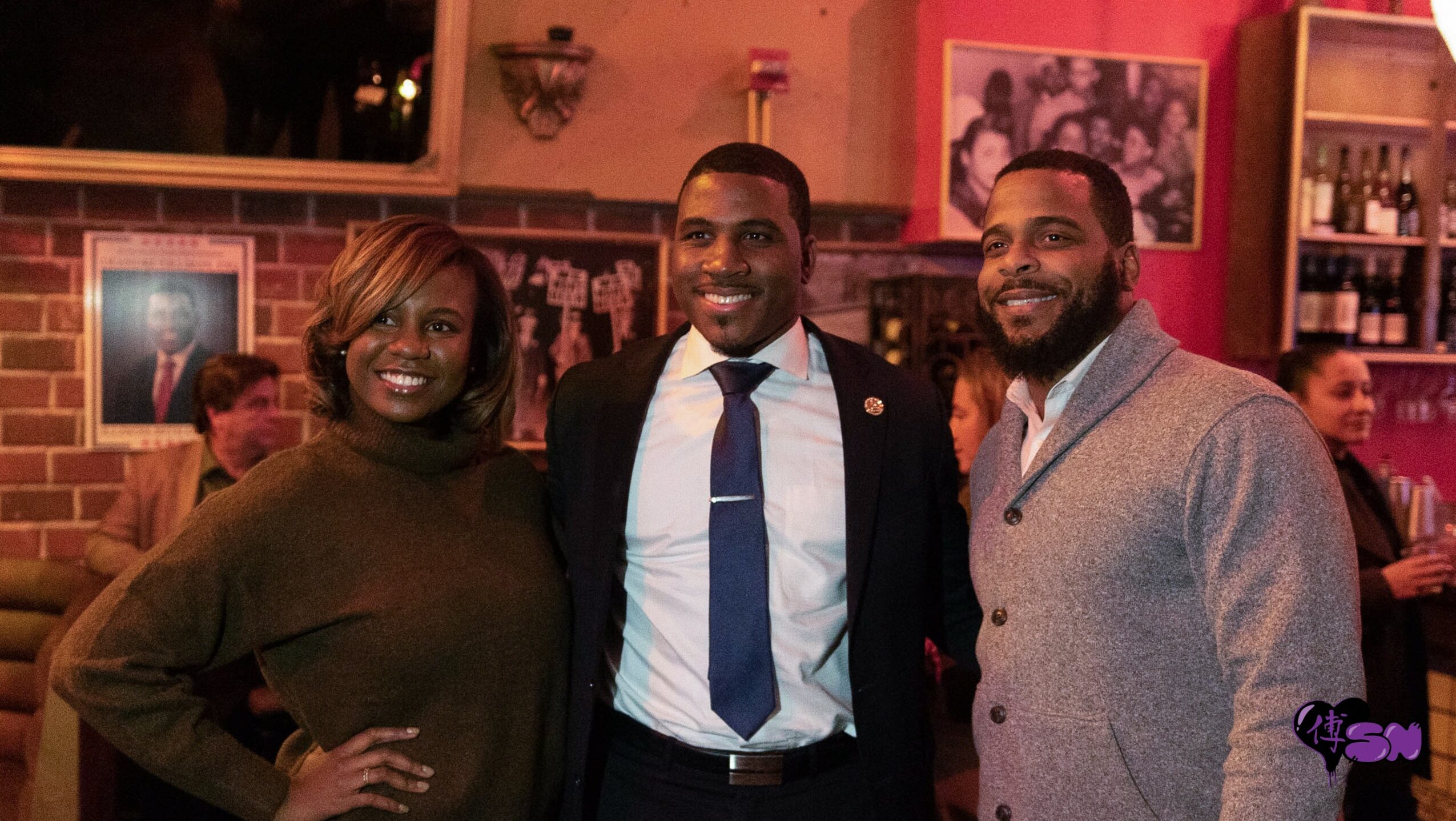 Three people smiling and posing together in a bar or restaurant setting with warm lighting and framed photos on the wall.