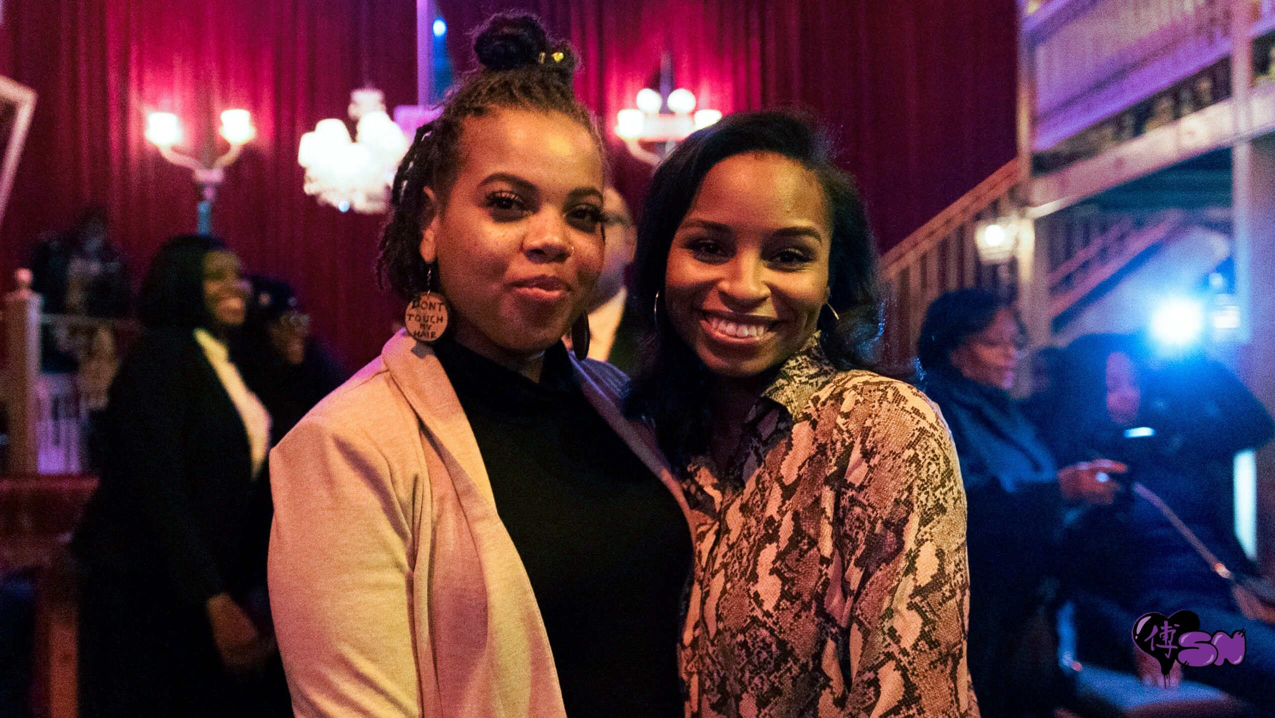 Two women smiling at a social event with red curtains and people in the background.