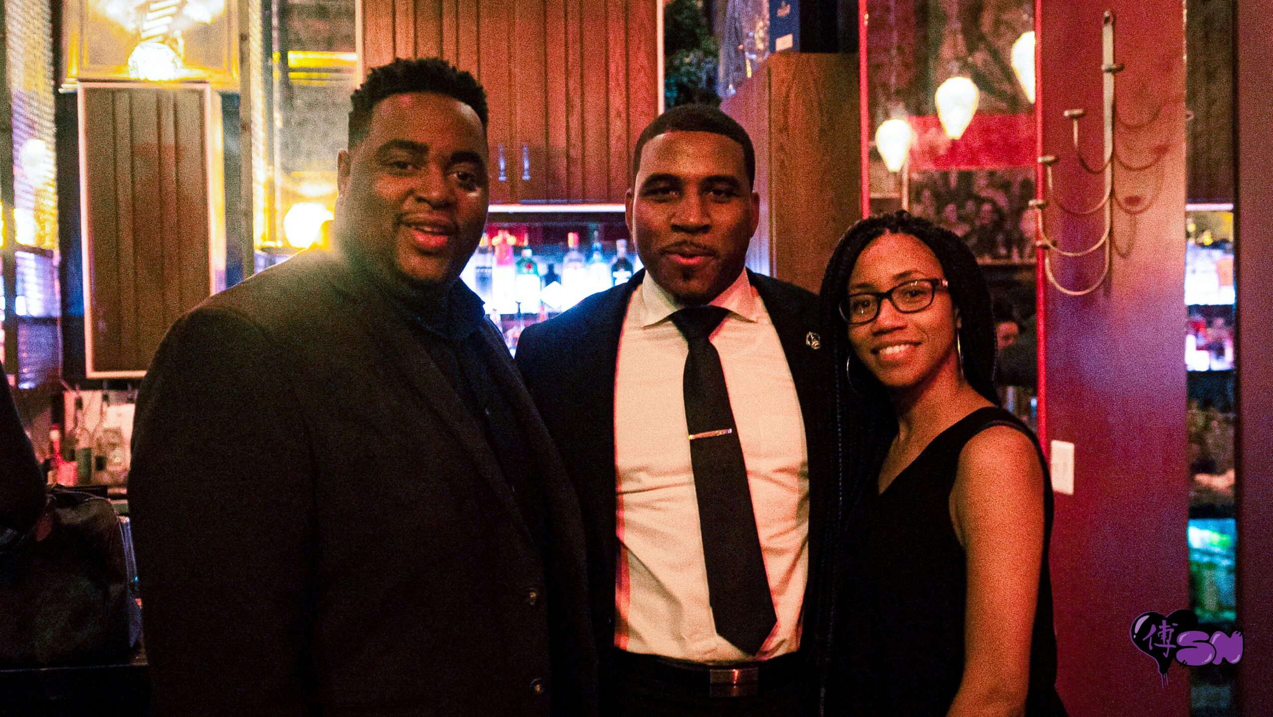 Three people standing together in a dimly lit indoor setting, smiling at the camera.