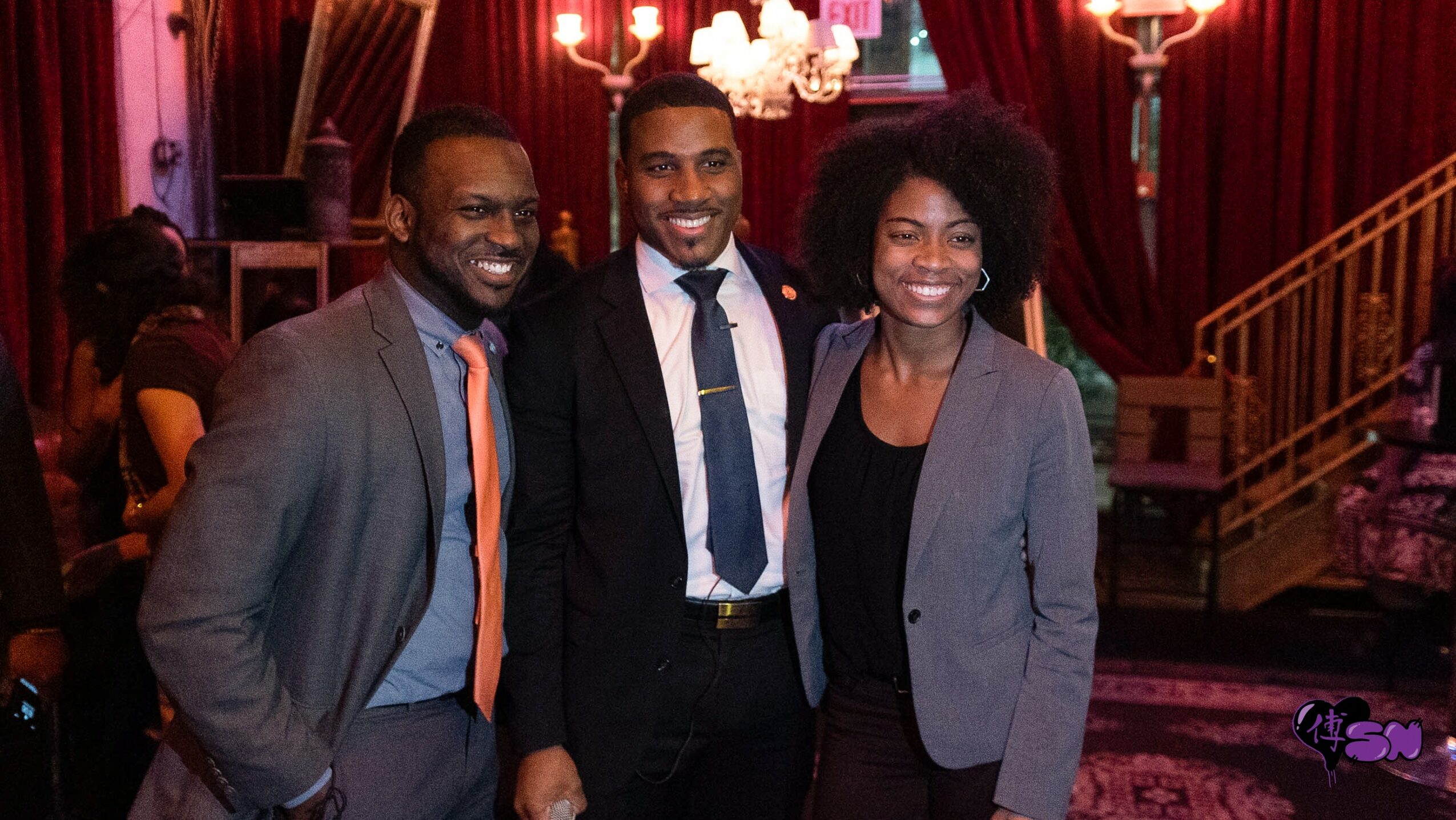 Three people in formal attire smiling indoors with red curtains and staircase in background.