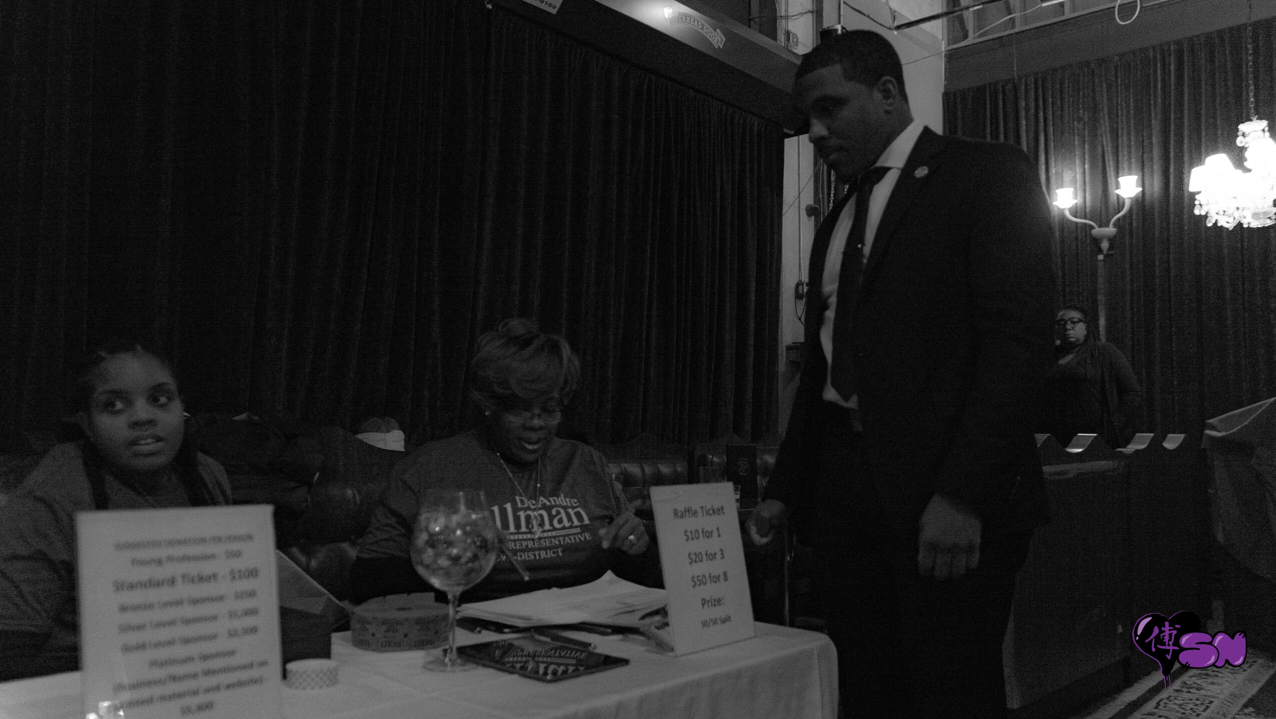 People sitting at a table with a man standing nearby in a dimly lit room, possibly at an event or gathering.
