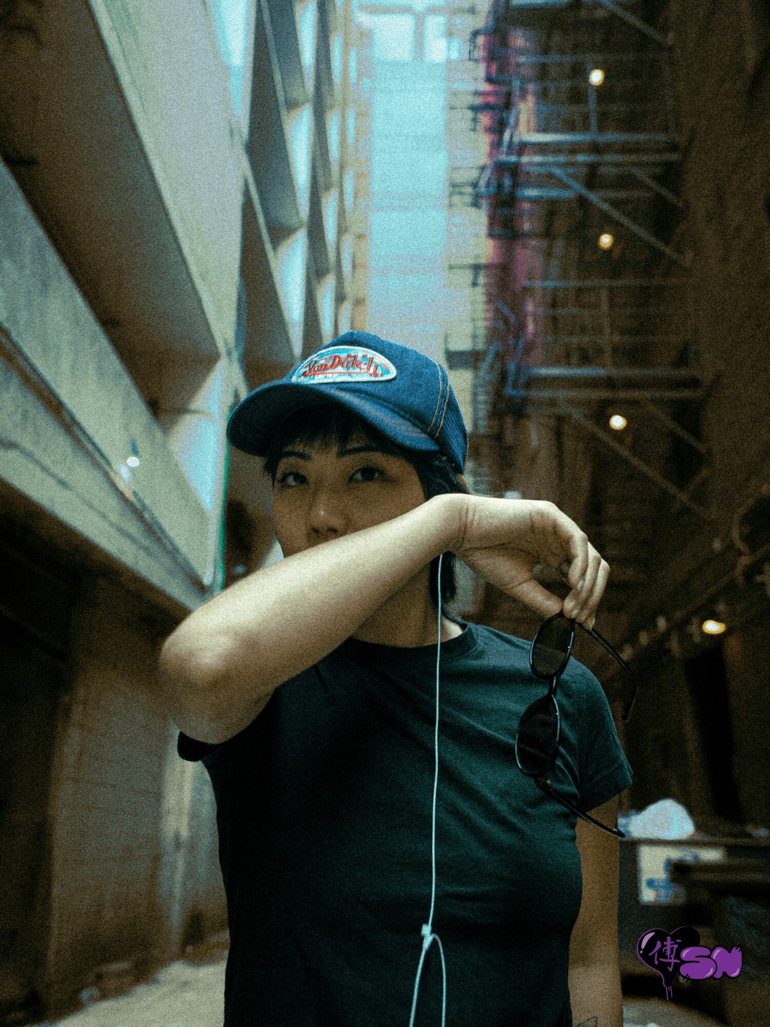 Person wearing a cap and black shirt, covering part of their face with their arm, in an indoor setting.