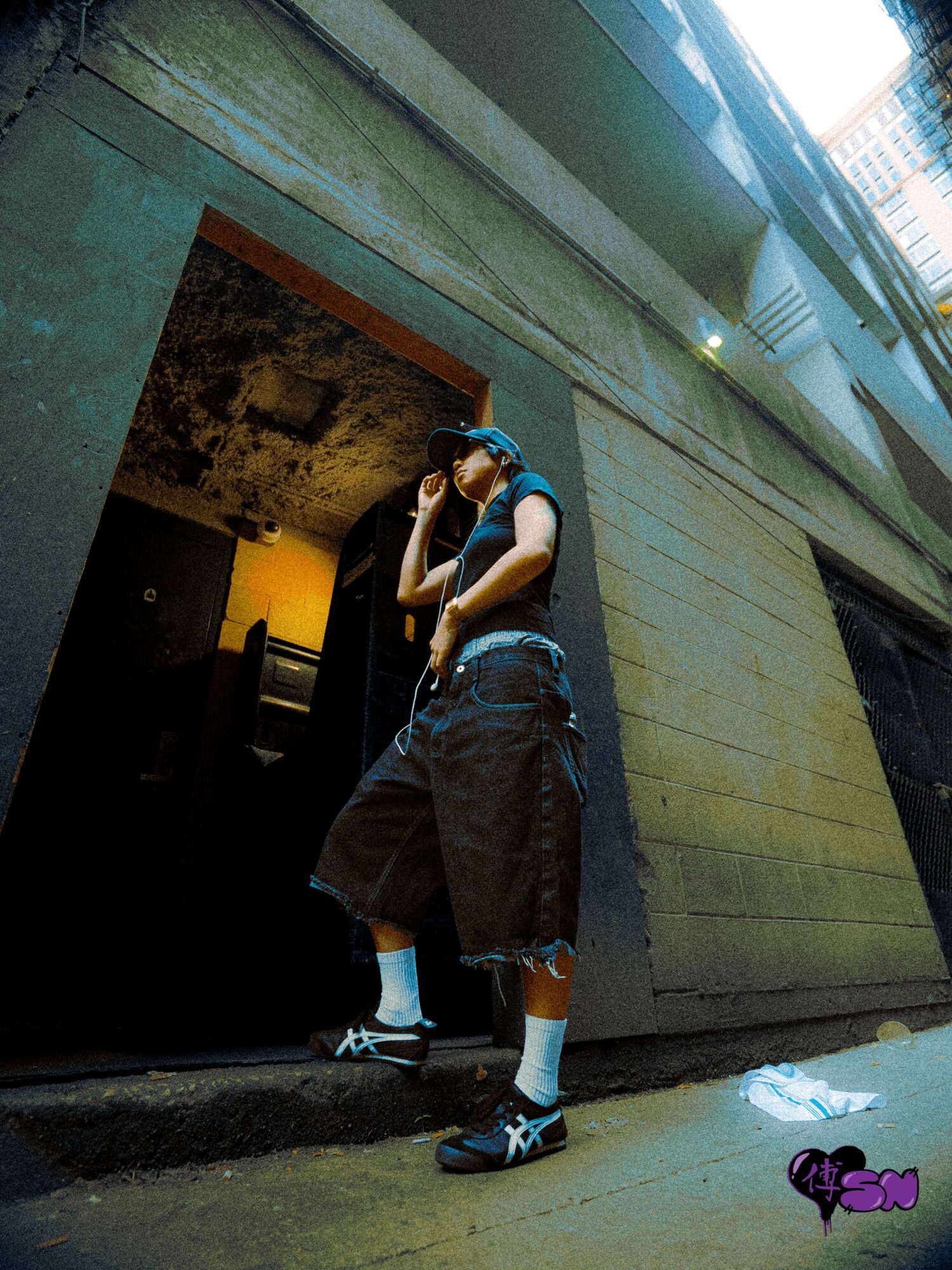 Person leaning against a wall in an urban setting, wearing a cap, baggy pants, and sneakers, with a building in the background.