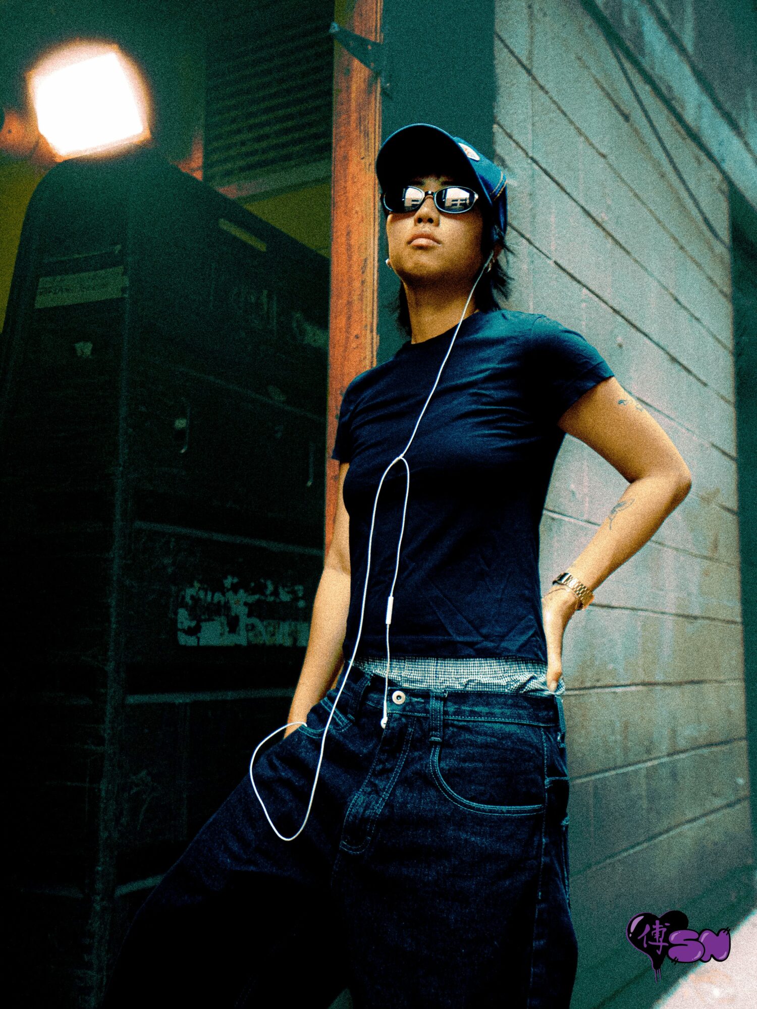 Woman wearing sunglasses and headphones, standing against a wall with her hand on her hip.