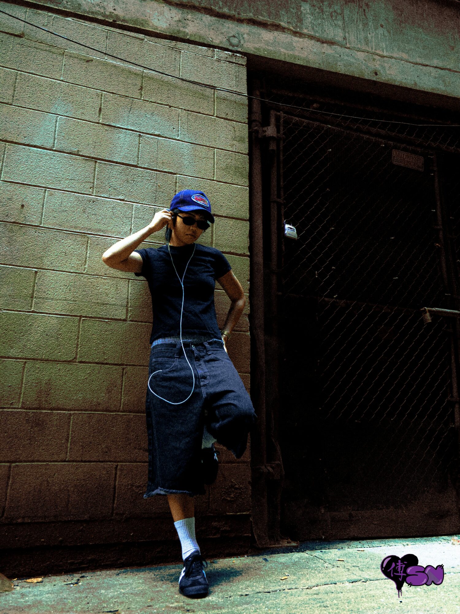 Person leaning against a brick wall, wearing a blue cap, sunglasses, black shirt, and cropped jeans, with one hand on their head.