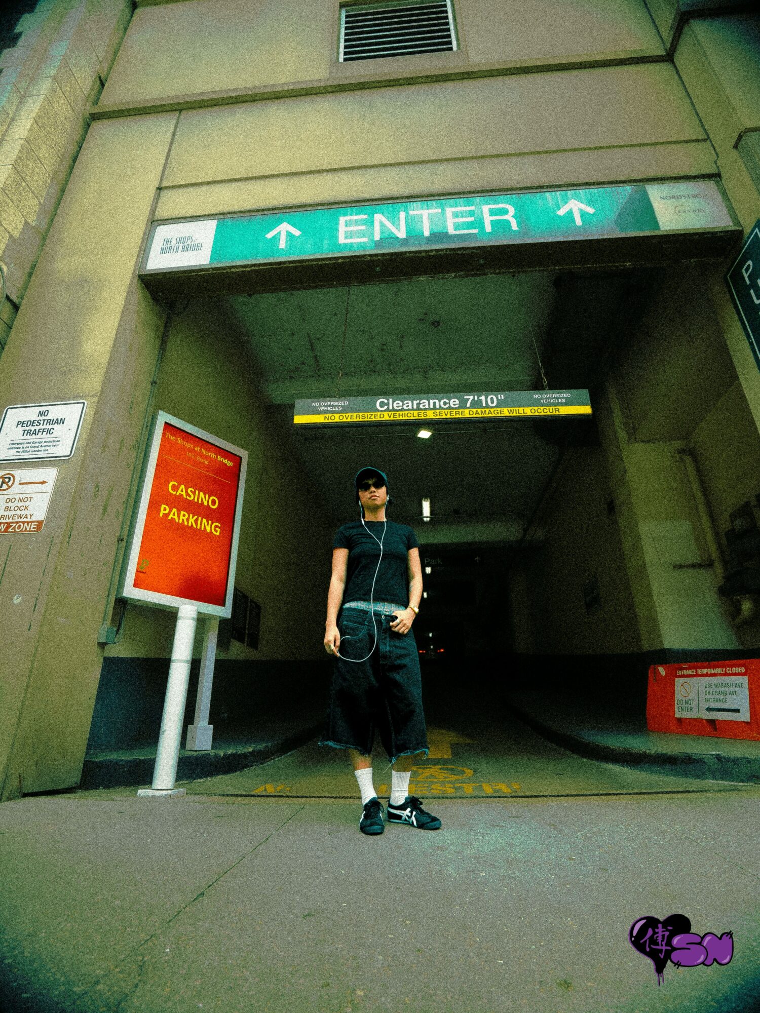 Person standing under an entrance sign with a parking garage in the background, wearing a black outfit and face mask.