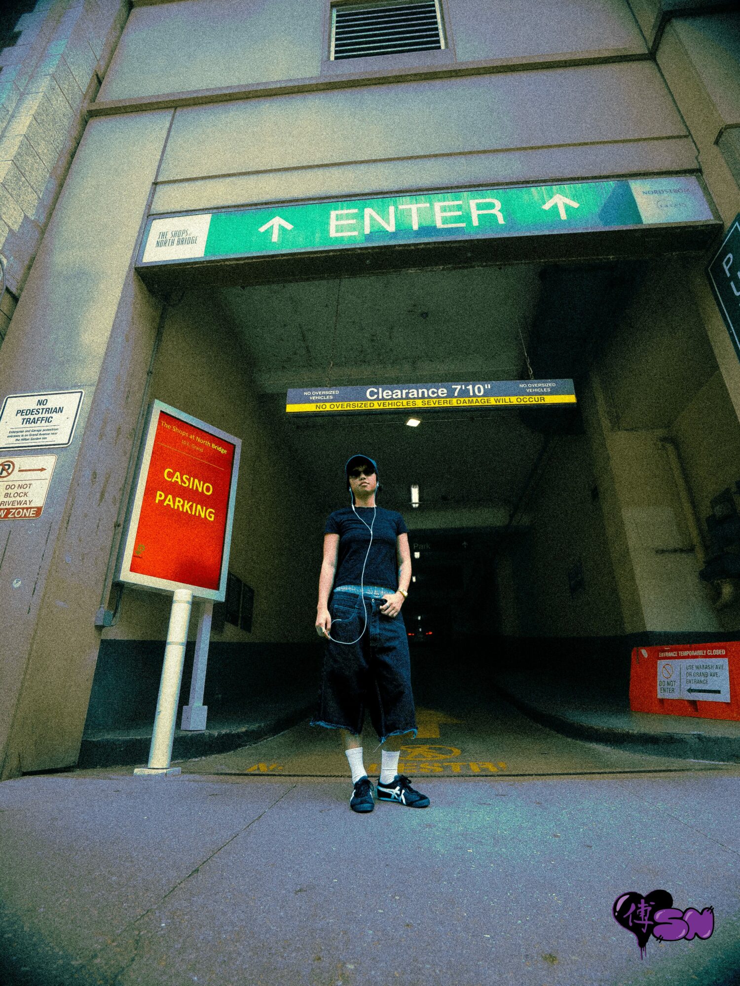 Person standing in front of parking garage entrance with green 'ENTER' sign above, wearing a mask, black shirt, and shorts.