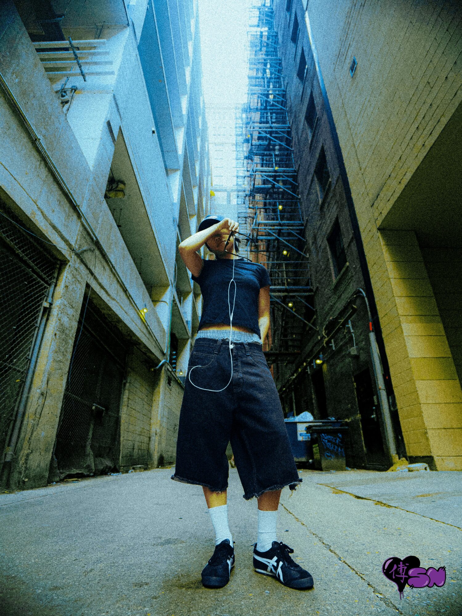 Young person standing in alleyway between tall buildings, looking up, wearing dark clothing, sneakers, and headphones, with scaffolding above.