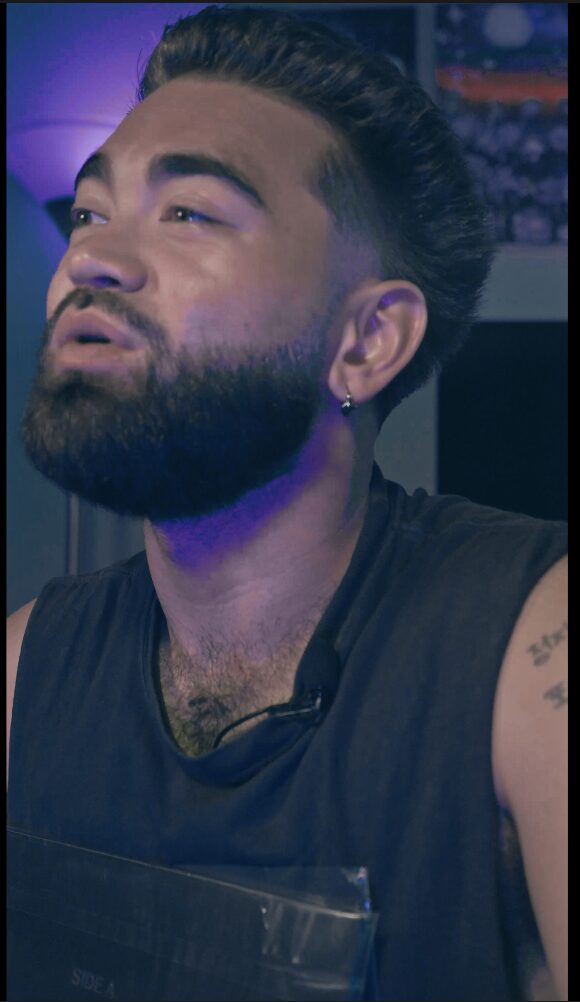 Man with dark hair and beard, wearing a sleeveless shirt, looking to the side, with a blurred background