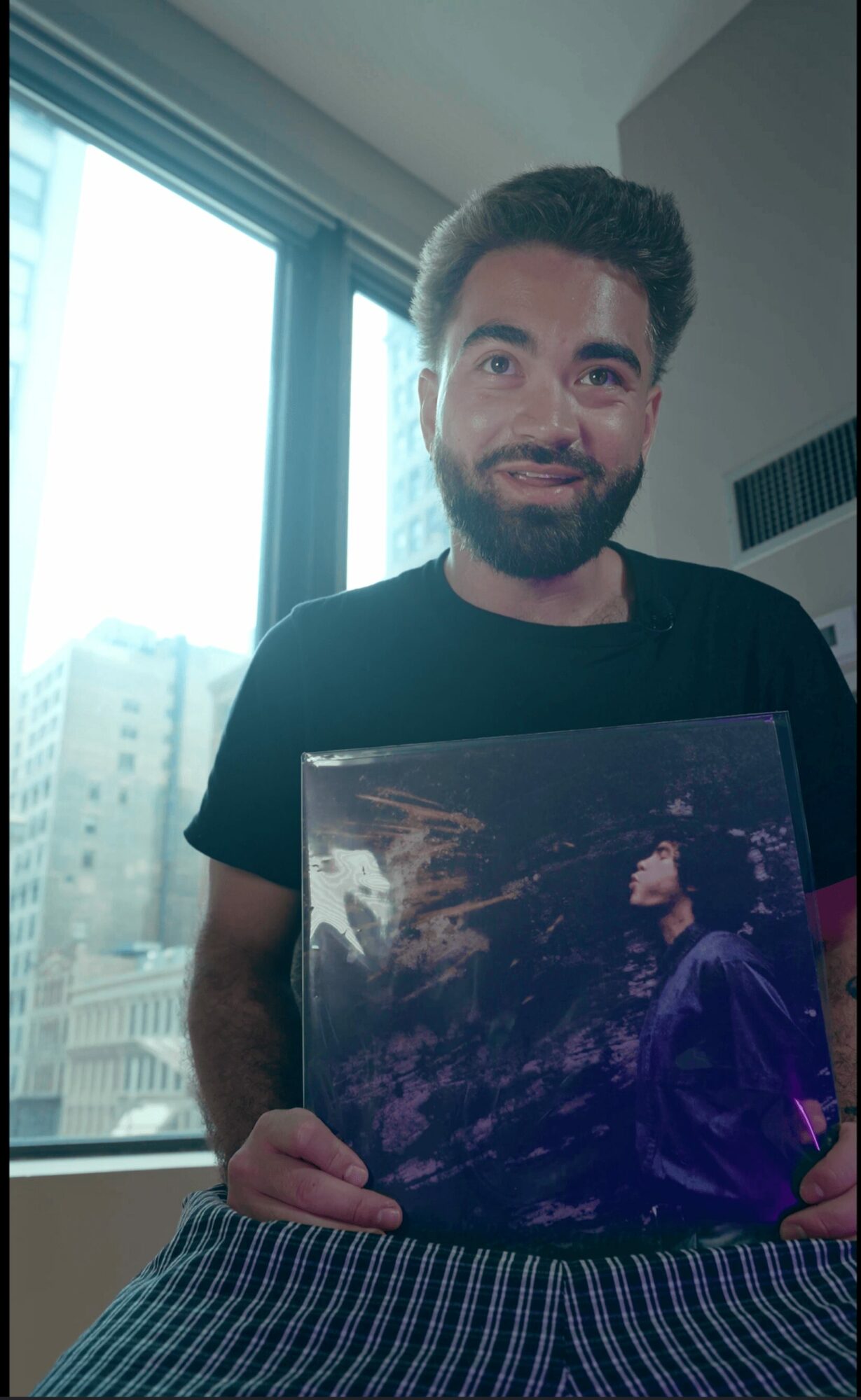 Young man with dark hair and beard holding a framed picture, standing indoors near a window with city buildings outside.